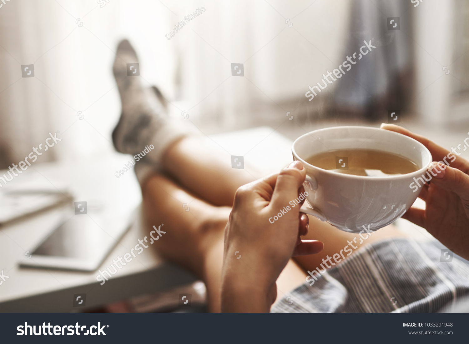Cup of tea and chill. Woman lying on couch, holding legs on coffee table, drinking hot coffee and enjoying morning, being in dreamy and relaxed mood. Girl in oversized shirt takes break at home #1033291948