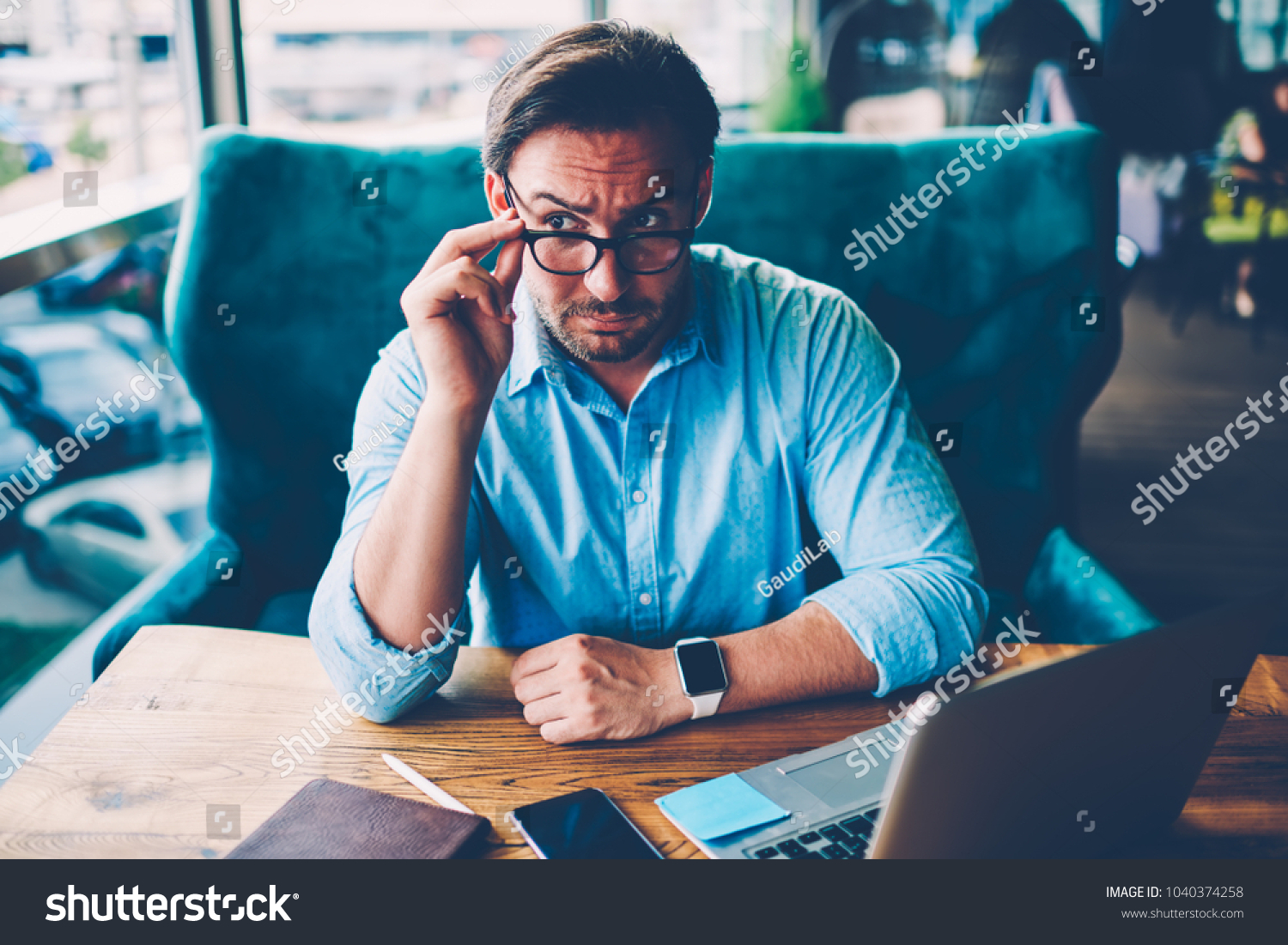 Bearded entrepreneur dressed in blue shirt fixing optical eyeglasses with black frame while looking away sitting at wooden desktop with digital laptop computer connected to wireless internet #1040374258