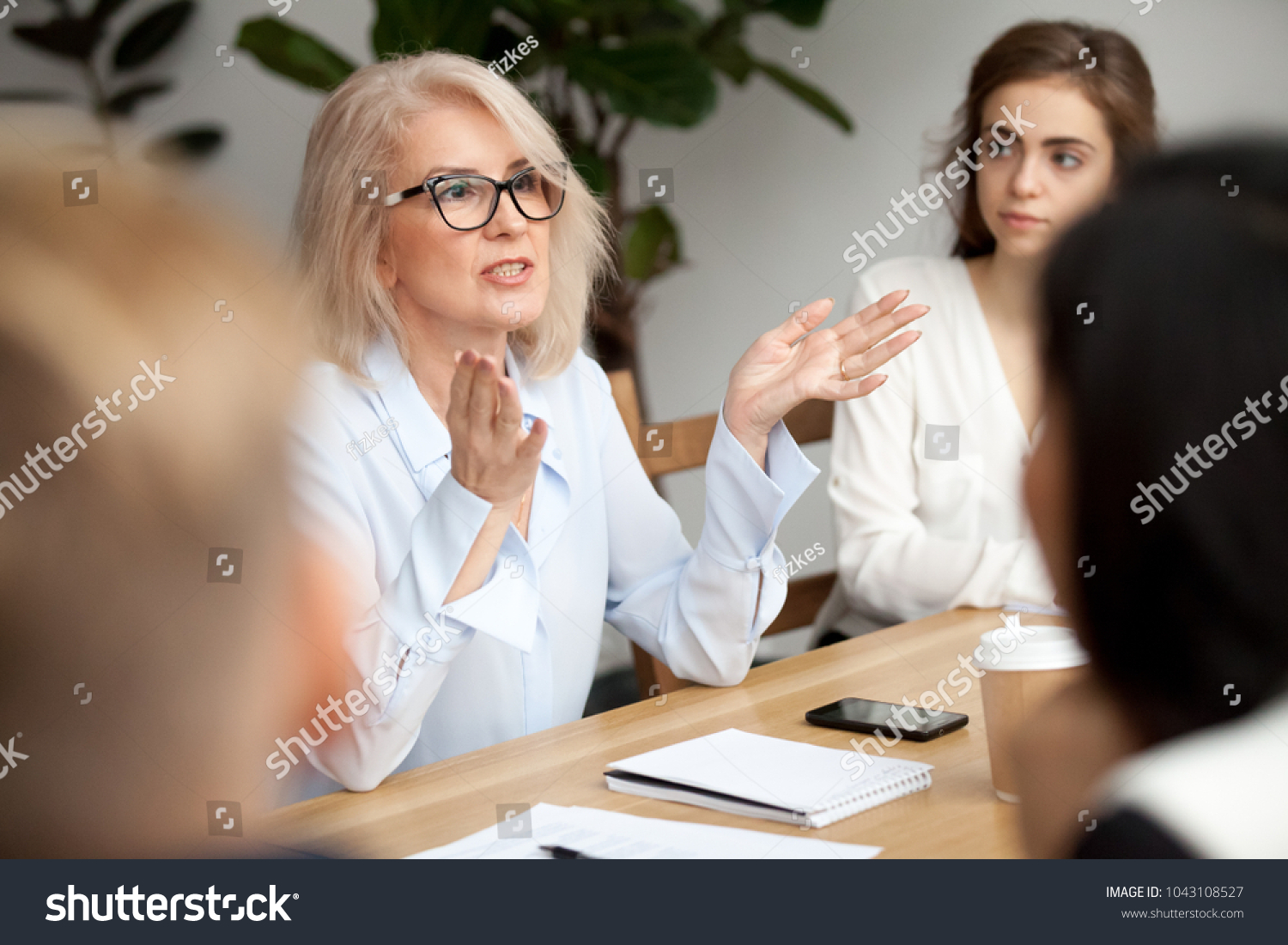 Attractive aged businesswoman, teacher or mentor coach speaking to young people, senior woman in glasses teaching audience at training seminar, female business leader speaker talking at meeting #1043108527