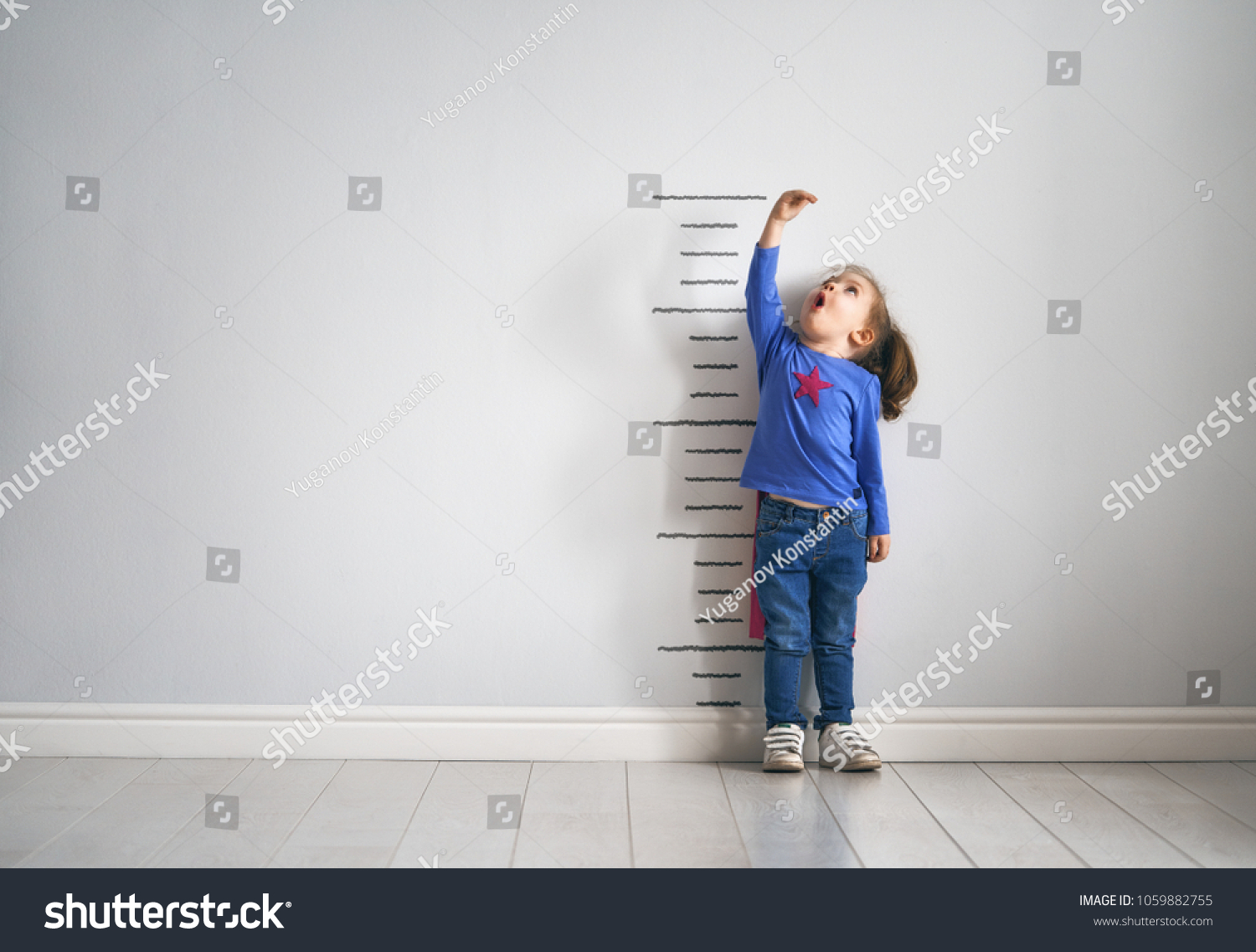 Little child is playing superhero. Kid is measuring the growth on the background of wall. Girl power concept.  #1059882755