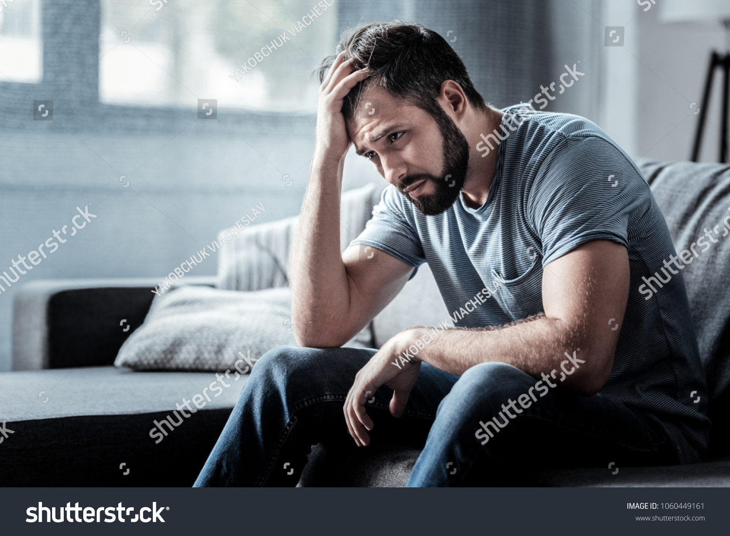 Unpleasant pain. Sad unhappy handsome man sitting on the sofa and holding his forehead while having headache #1060449161