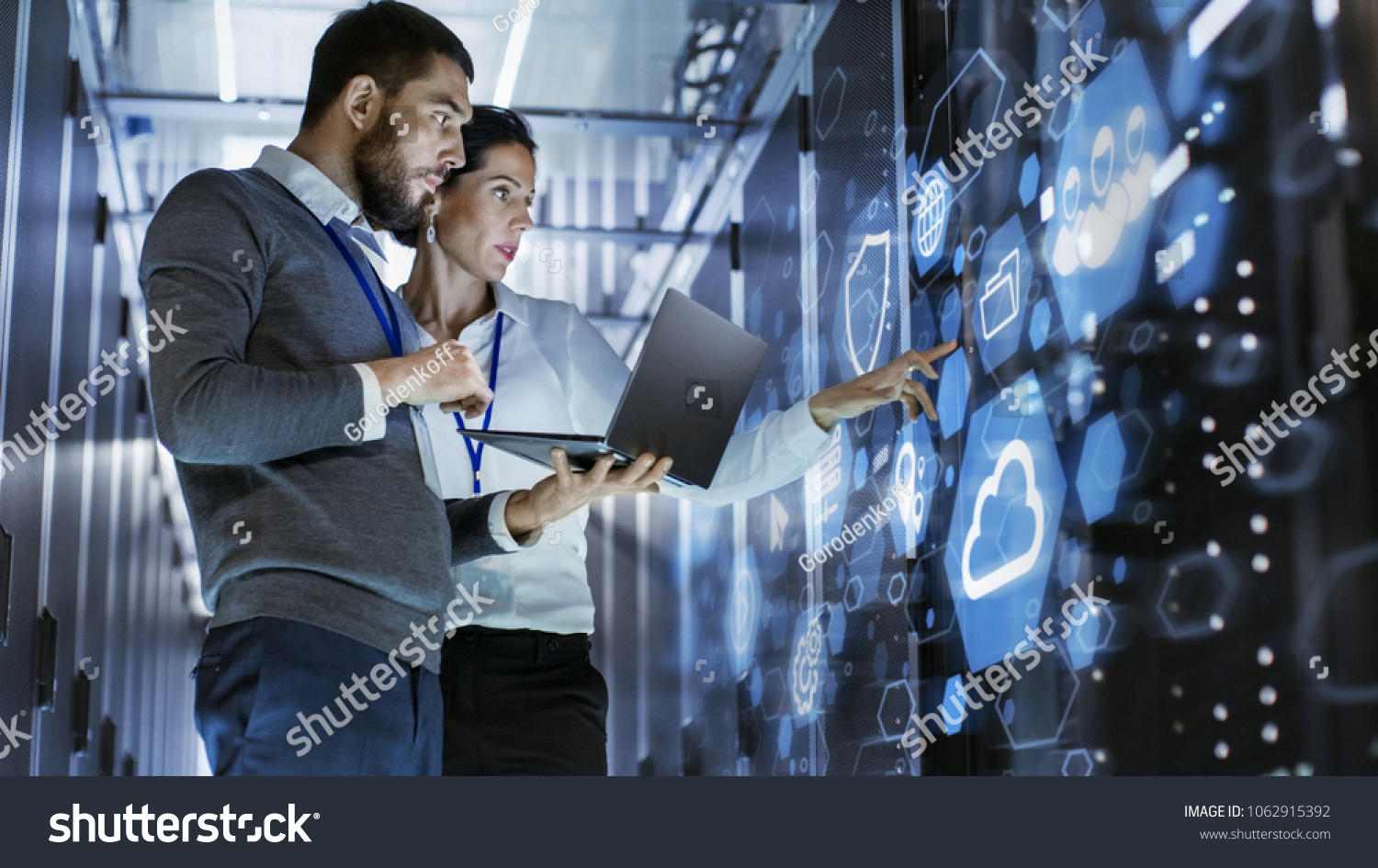 Der männliche IT-Spezialist hält Laptop und diskutiert die Arbeit mit weiblichen Servertechnikern. Sie stehen im Rechenzentrum, im Rack-Serverschrank mit Cloud-Server-Symbol und Visualisierung. #1062915392