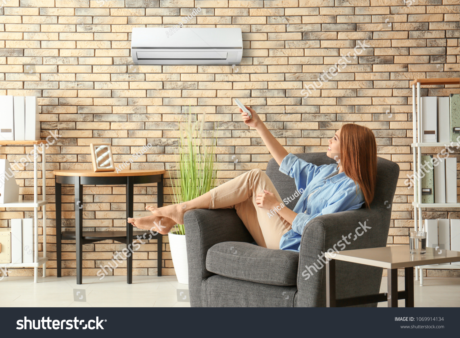 Young woman switching on air conditioner while sitting in armchair at home #1069914134