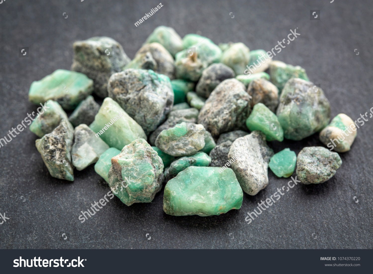 a pile of raw emerald gemstones (mineral beryl) with inclusions mined in Brazil on a gray slate stone #1074370220