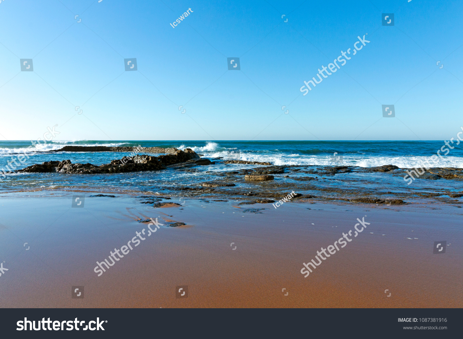 Wet smooth shoreline beach sand with rocks ocean and waves against blue coastal skyline in Kwa-Zulu Natal, South Africa #1087381916