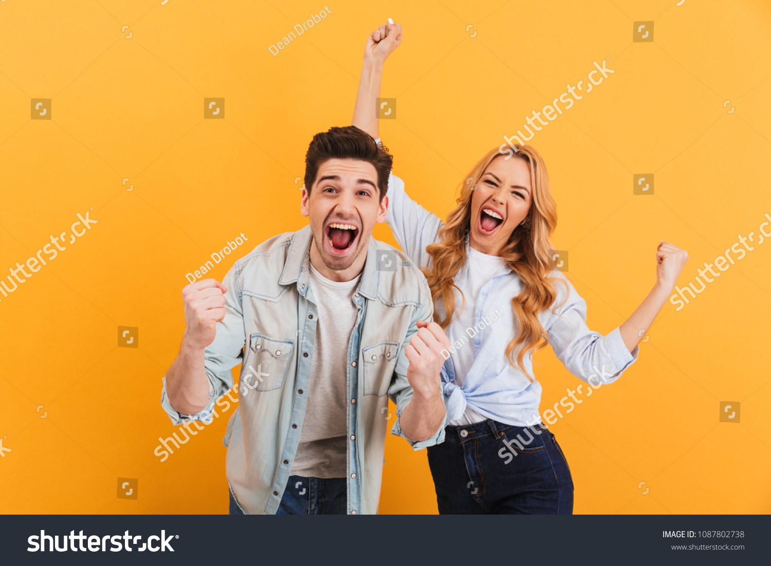 Portrait of cheerful people man and woman in basic clothing smiling and clenching fists like winners or happy people isolated over yellow background #1087802738