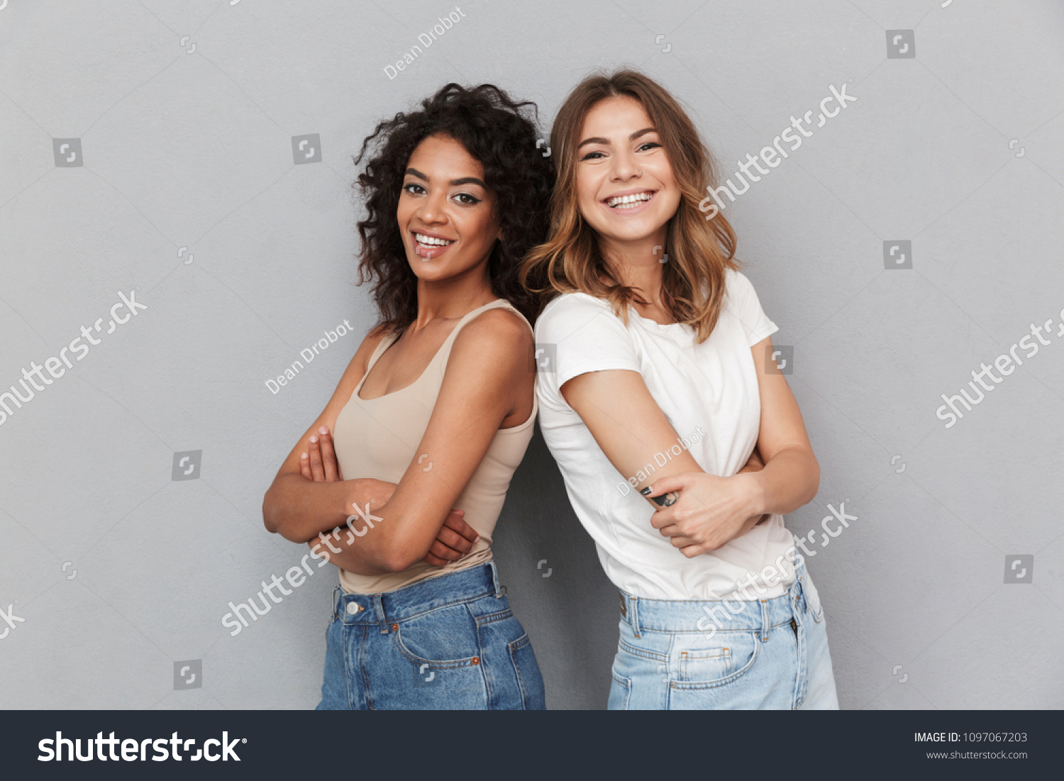 Portrait of two cheerful young women standing together and looking at camera isolated over gray background #1097067203