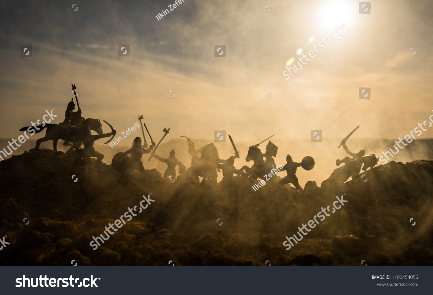 Medieval battle scene with cavalry and infantry. Silhouettes of figures as separate objects, fight between warriors on sunset foggy background. Selective focus #1100454056