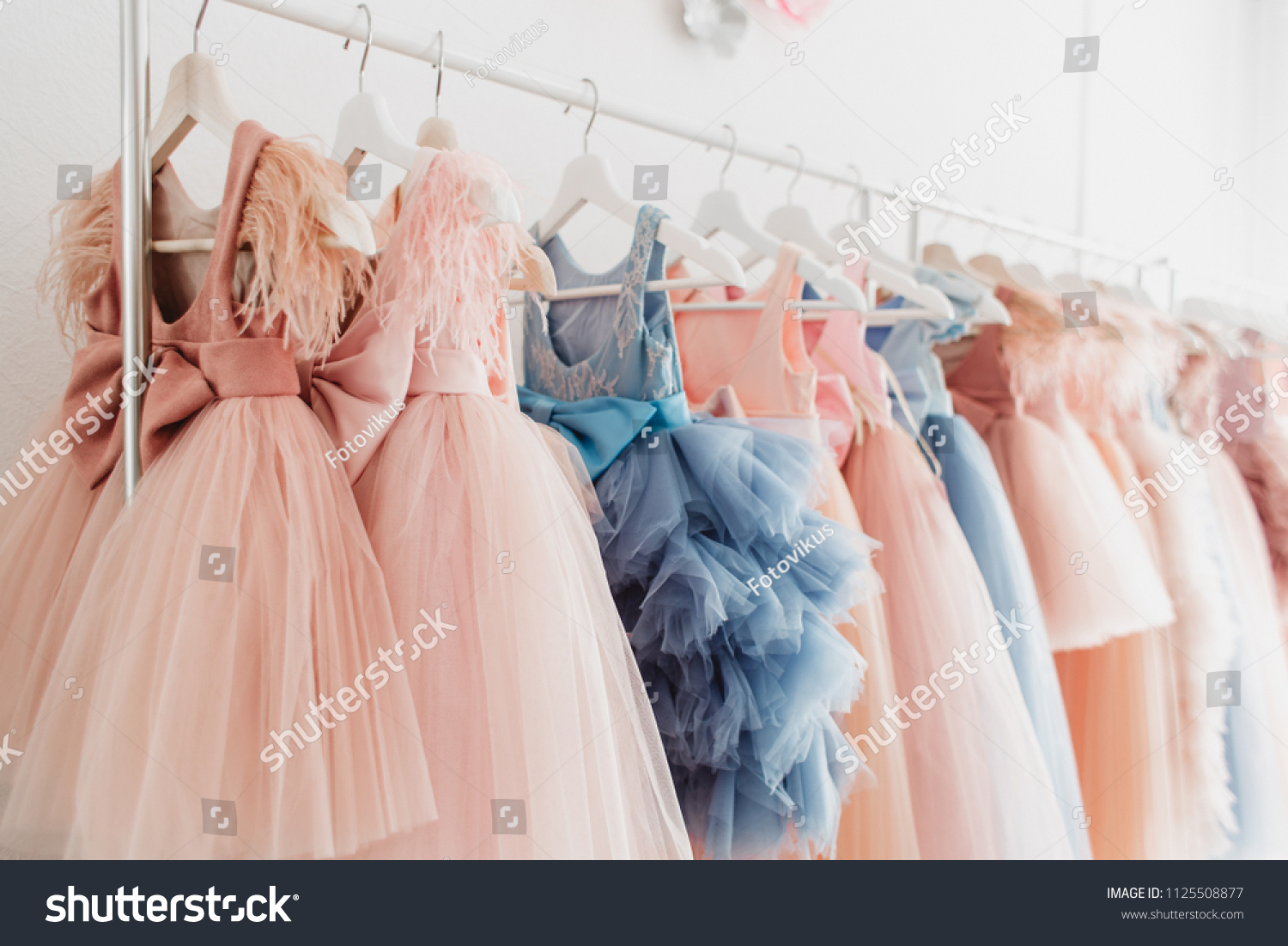 Beautiful dressy lush pink and blue dresses for girls on hangers at the background of white wall. Kids dresses with feathers for prom and holiday. #1125508877