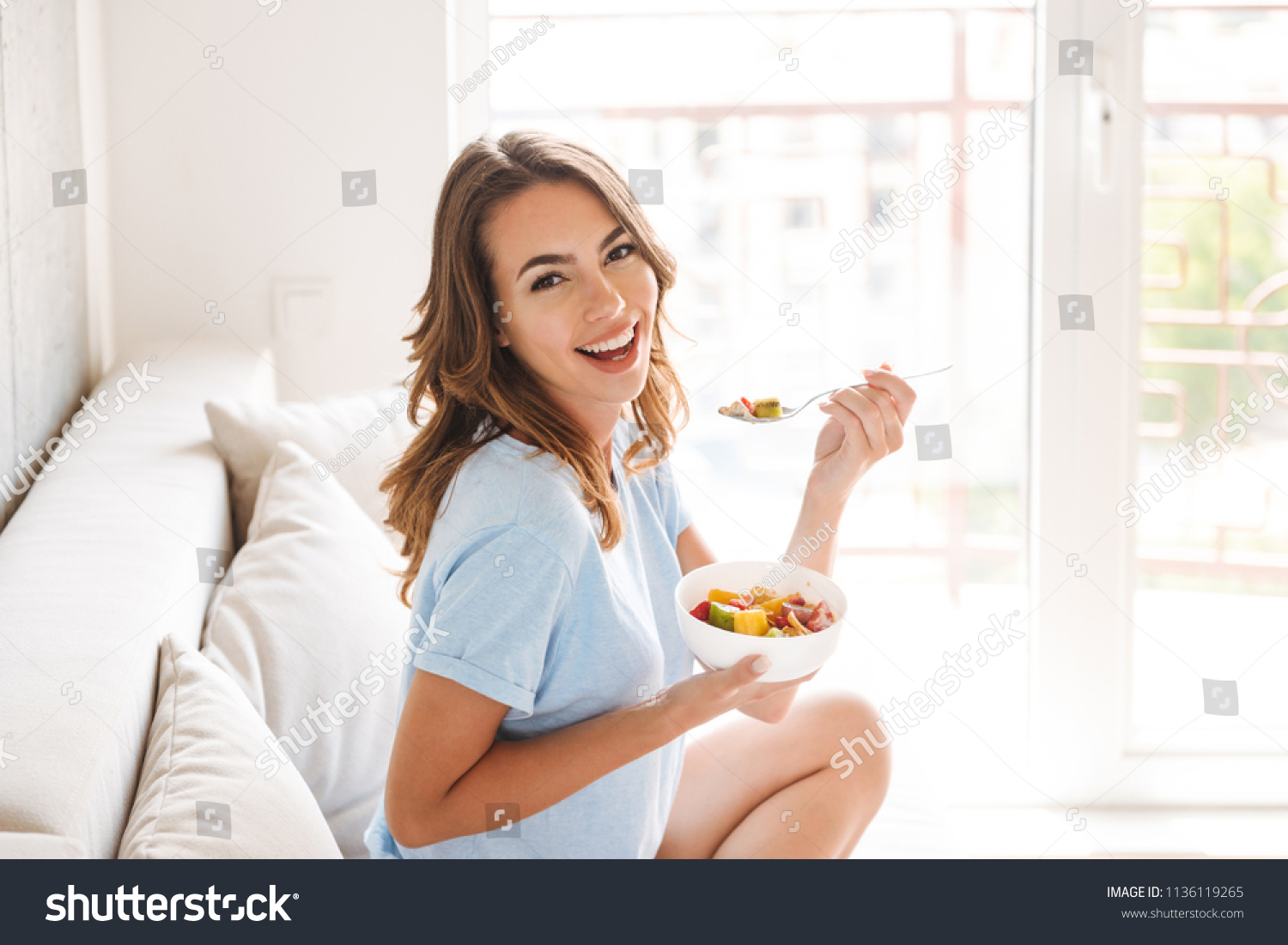 Cheerful young woman eating healthy breakfast while sitting on a couch at home #1136119265