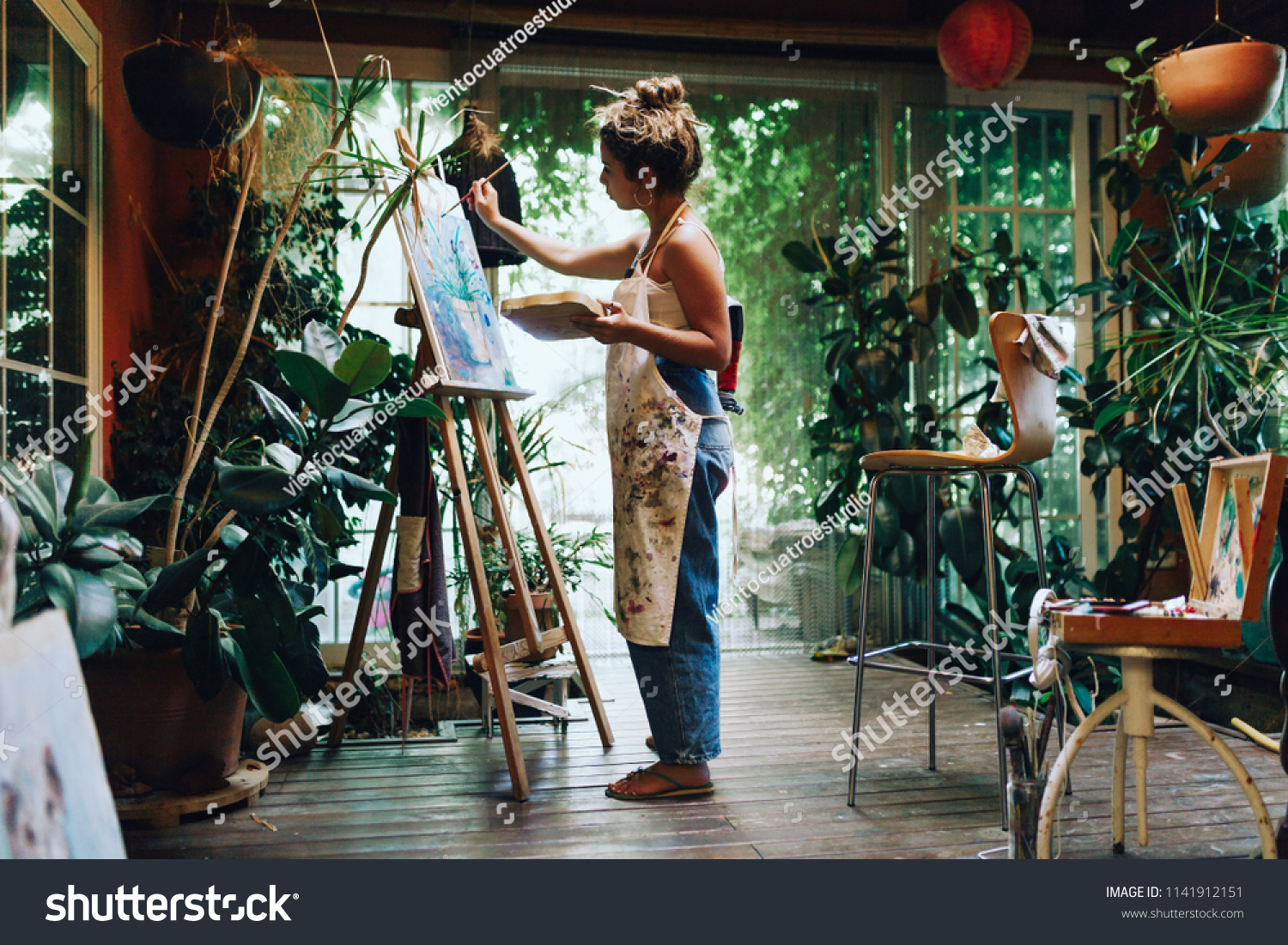 Indoor shot of professional female artist painting on canvas in studio with plants. #1141912151