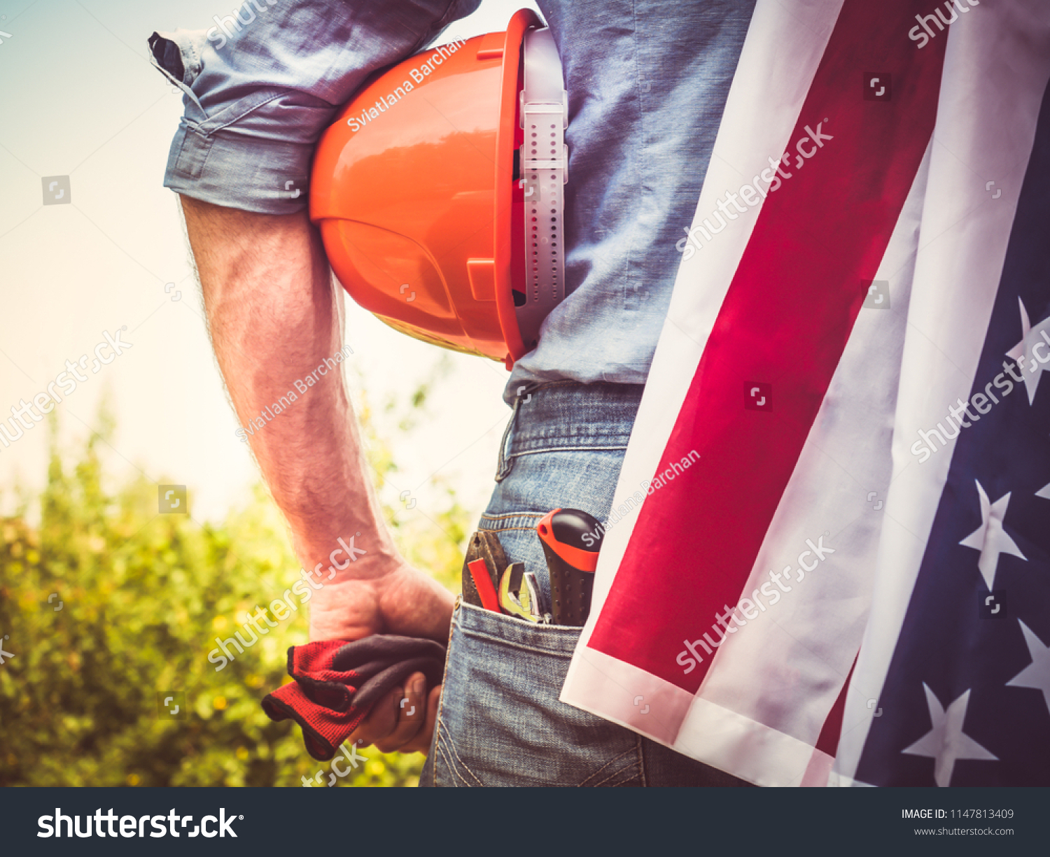 Attractive man in work clothes, holding tools and a US flag in his hands and looking into the distance against the background of trees, blue sky and sunset. View from the back. Labour Day Concept #1147813409