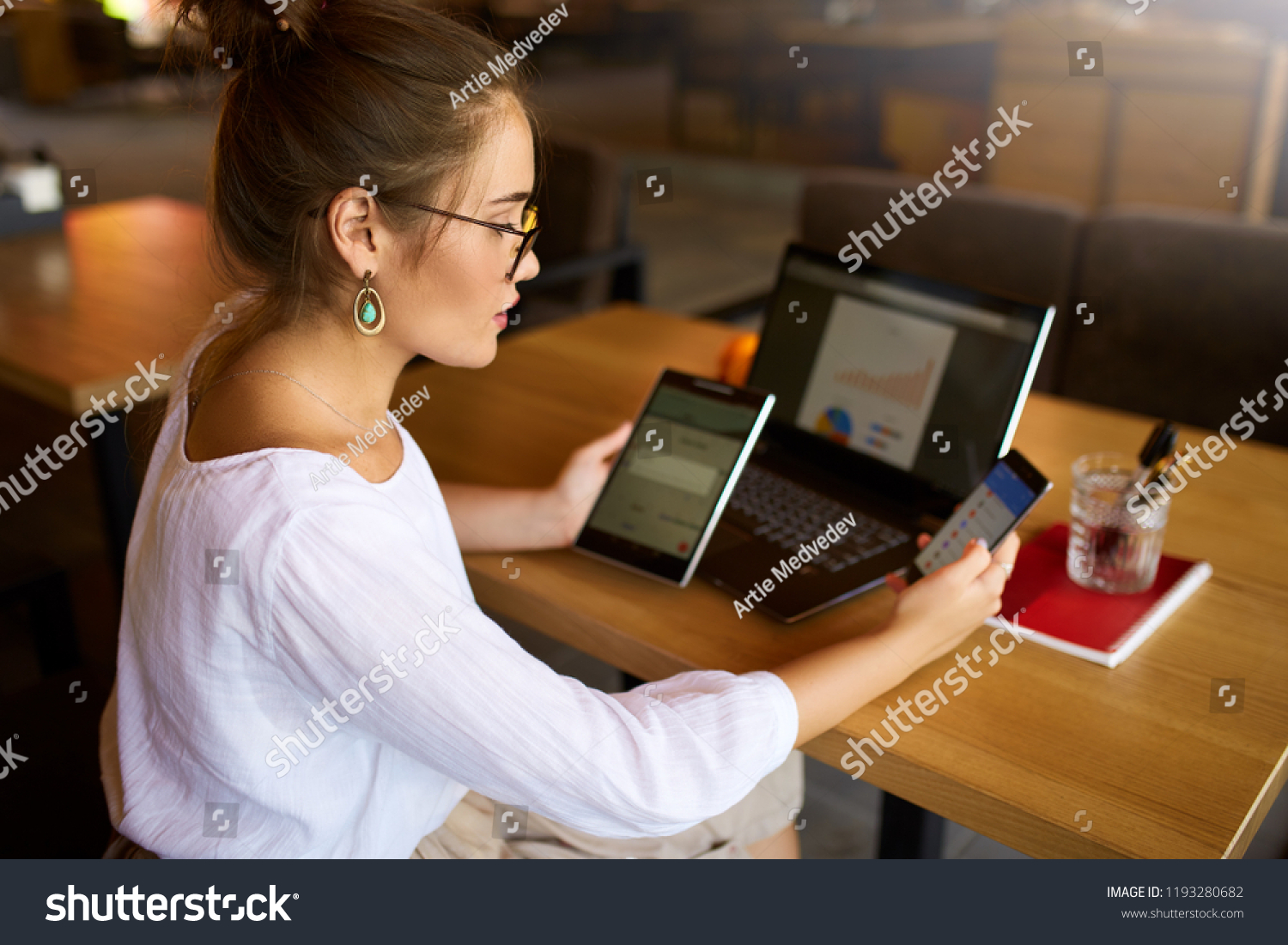 Mixed race woman in glasses working with multiple electronic internet devices. Freelancer businesswoman has tablet and cellphone in hands and laptop on table with charts on screen. Multitasking theme #1193280682