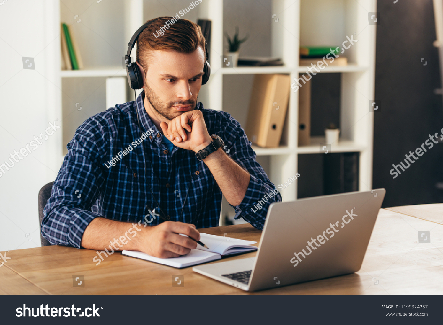 portrait of concentrated man in headphones taking part in webinar at tabletop with notebook in office #1199324257