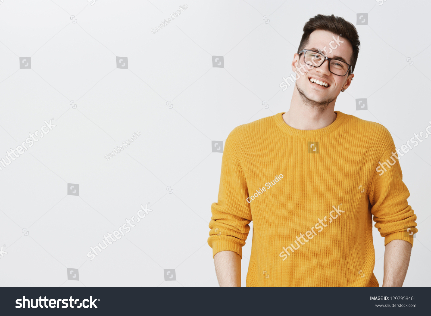 Waist-up shot of happy and delighted handsome young man in glasses and yellow sweater tilting head, smiling and laughing as looking friendly at camera on right side of copy space over gray background #1207958461