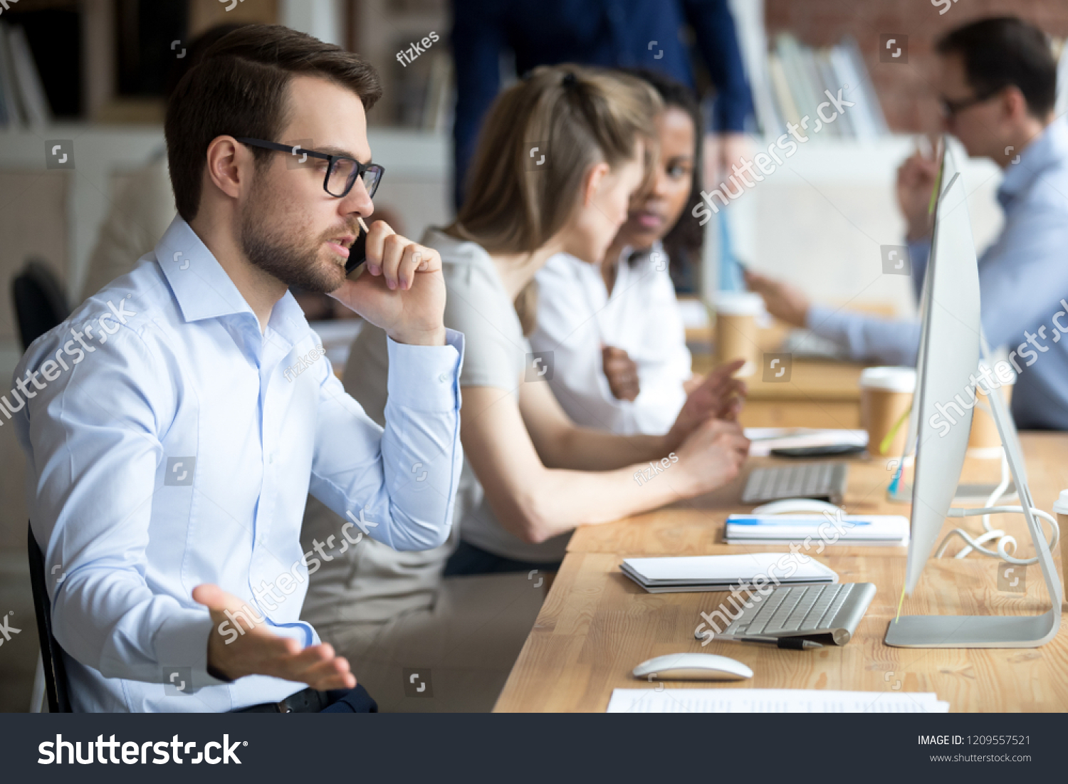 Angry male employee confused working at computer, solve business problems on phone, mad worker talk on cell arguing with client or customer, man busy at pc having serious conversation on cellphone #1209557521