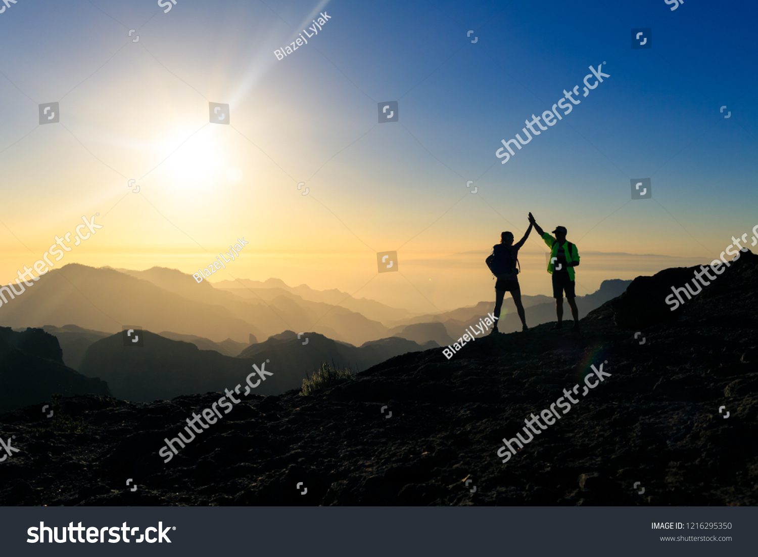 Couple hikers celebrating success in sunset mountains, accomplish with arms up outstretched. Young man and woman looking at beautiful inspirational landscape view, Gran Canaria Canary Islands. #1216295350