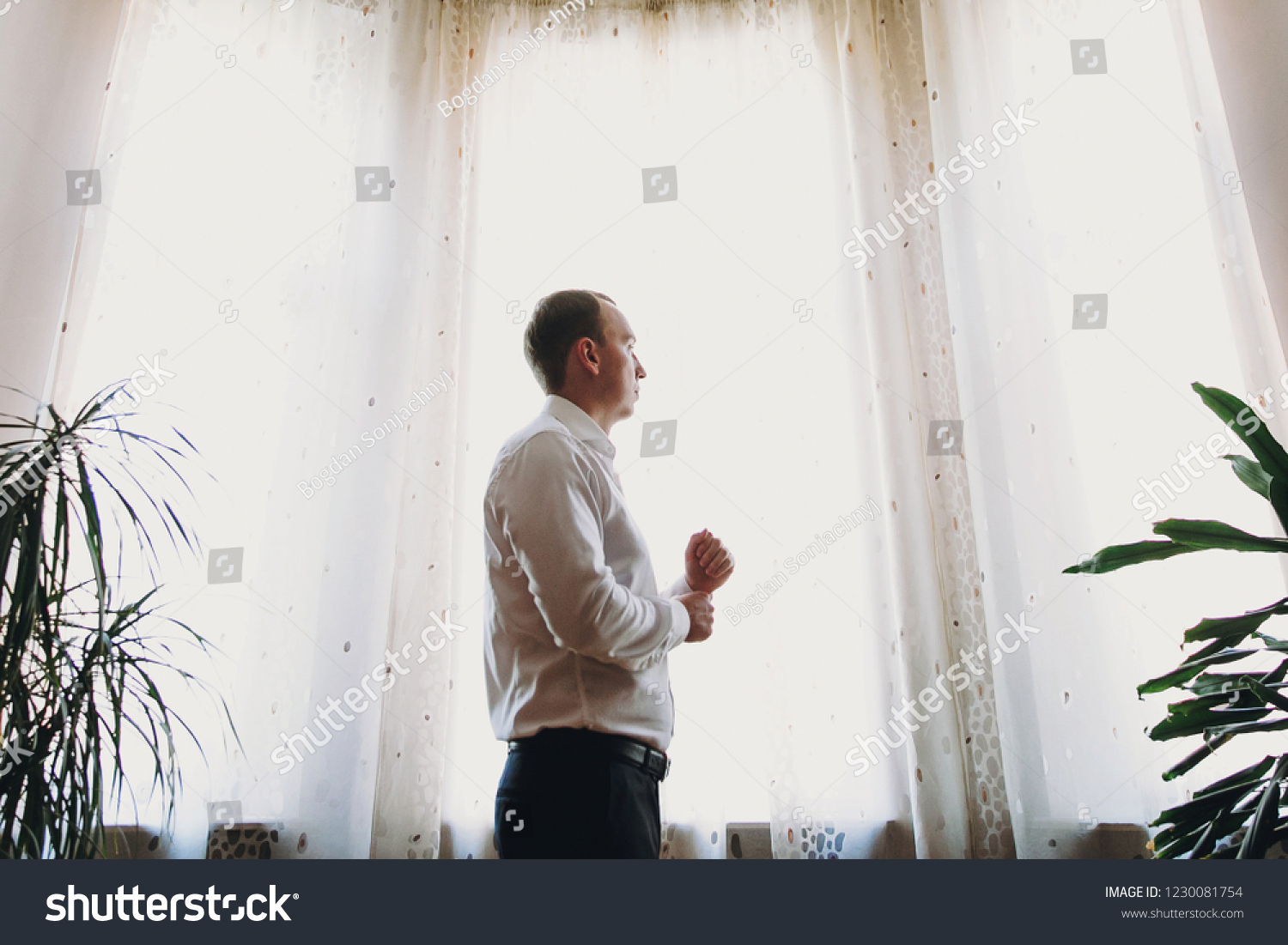 Stylish groom putting on white shirt, getting ready in the morning near window in soft light, before wedding ceremony #1230081754