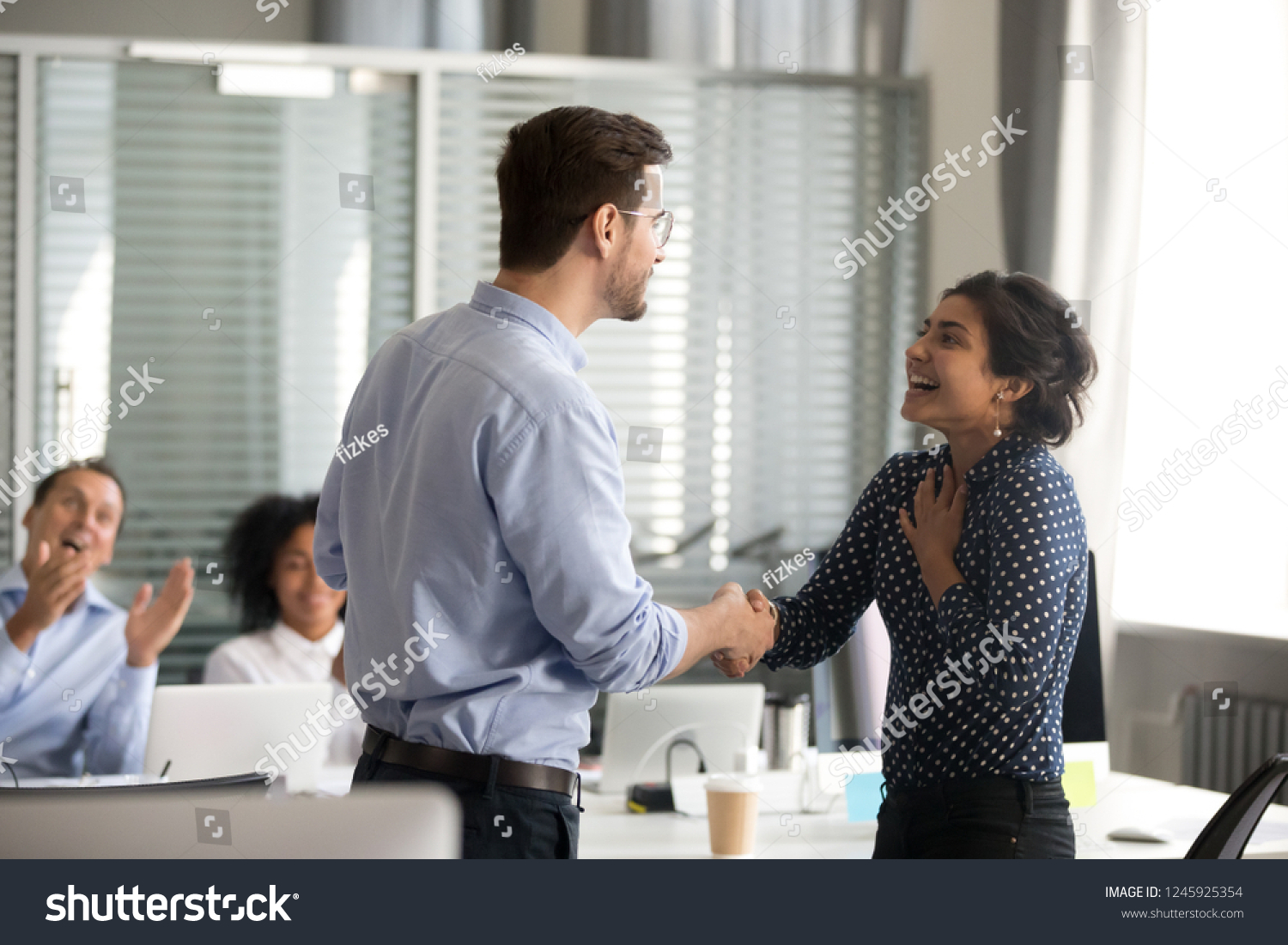 Team leader motivating handshaking female Indian excited employee, congratulate with promotion, business achievement, thank for good result, work, expressing respect, support, colleagues applaud #1245925354