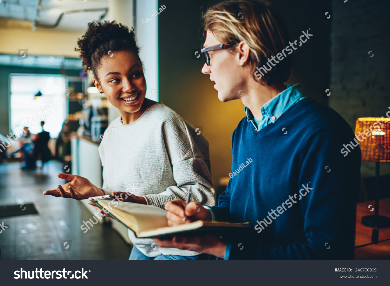 Positive multiracial male and female students having conversation about common project in cafe making notes, man writing to do list in notepad while talking and discussing with his girlfriend #1246756909