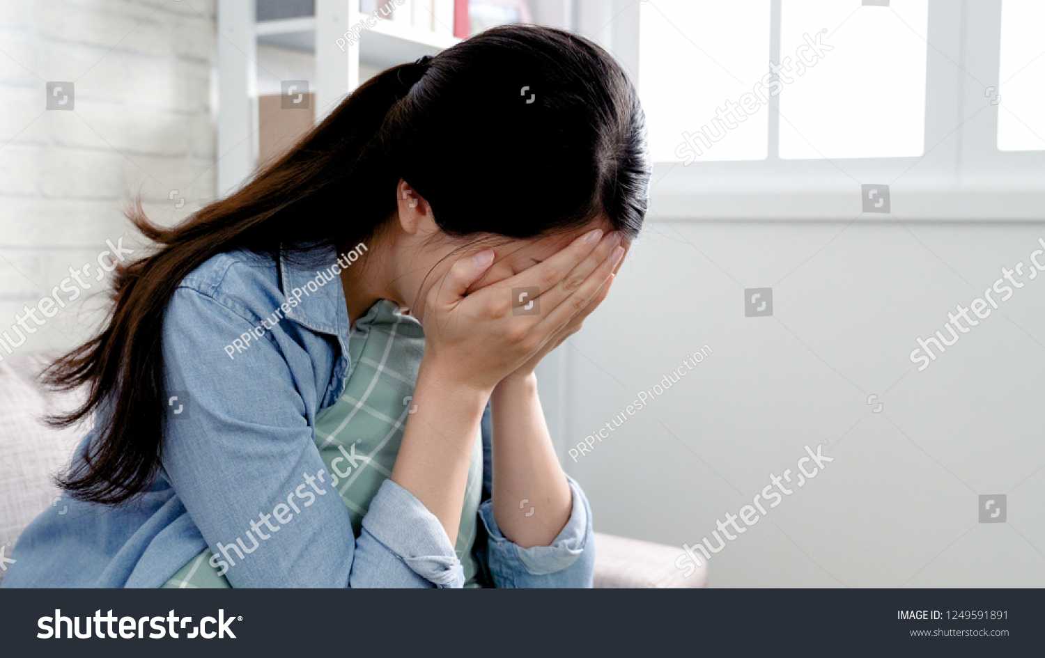 asian woman in cozy room sitting in sofa using hands cover face disappointed crying. emotional girl lovelorn tears heart pain concept staying home feeling broken. unhappy lady sadness indoors. #1249591891