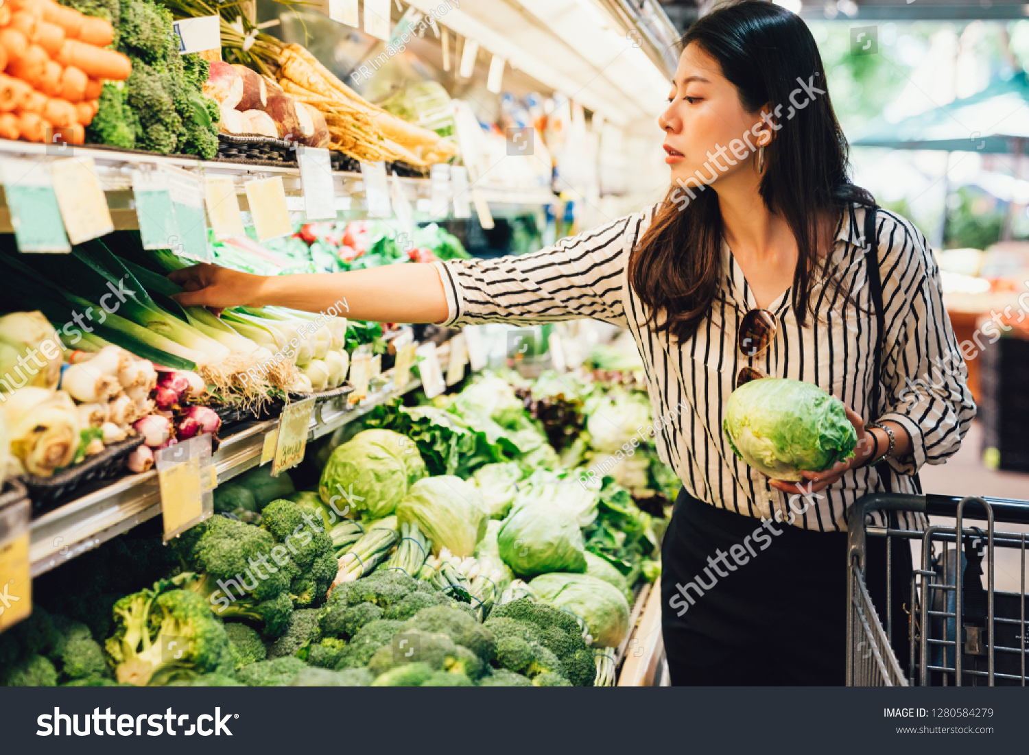 asian local woman buy vegetables and fruits in supermarket. young chinese lady holding green leaf vegetable and picking choosing green onion on cold open refrigerator. elegant female grocery shopping #1280584279