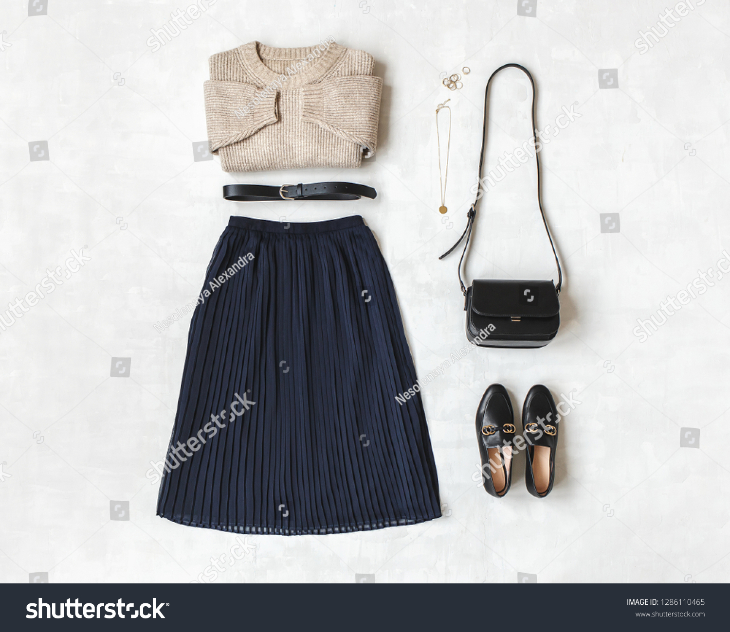 Blue midi pleated skirt, beige knitted sweater, small black cross body bag, belt, loafers (flat shoes) on grey background. Overhead view of women's casual day outfit. Flat lay, top view. Women clothes #1286110465