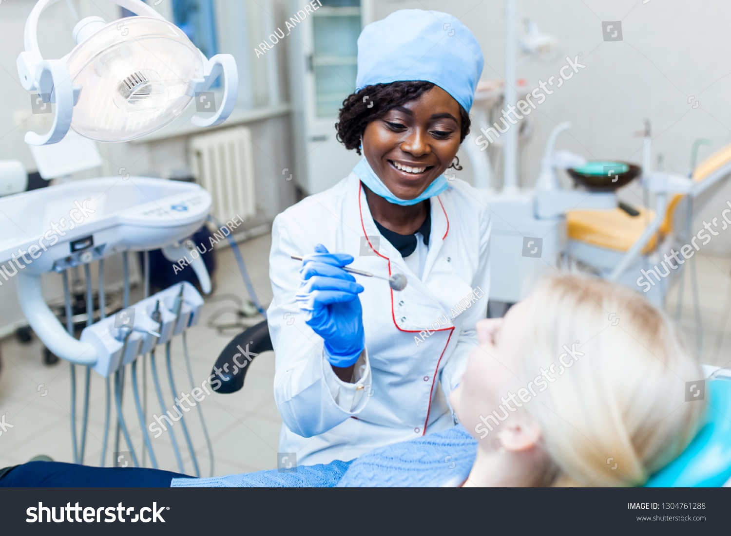 Female black dentist in dental office talking with female patient and preparing for treatment. Modern medical equipment. Dental clinic concept #1304761288