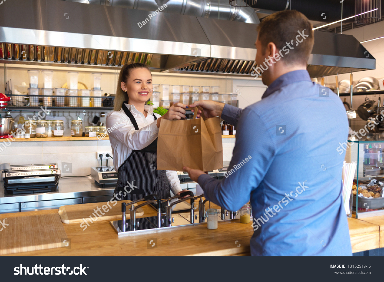 Cheerful waitress wearing apron serving customer at counter in restaurant - Small business and service concept with young business owner woman giving bag with takeaway food to client #1315291946