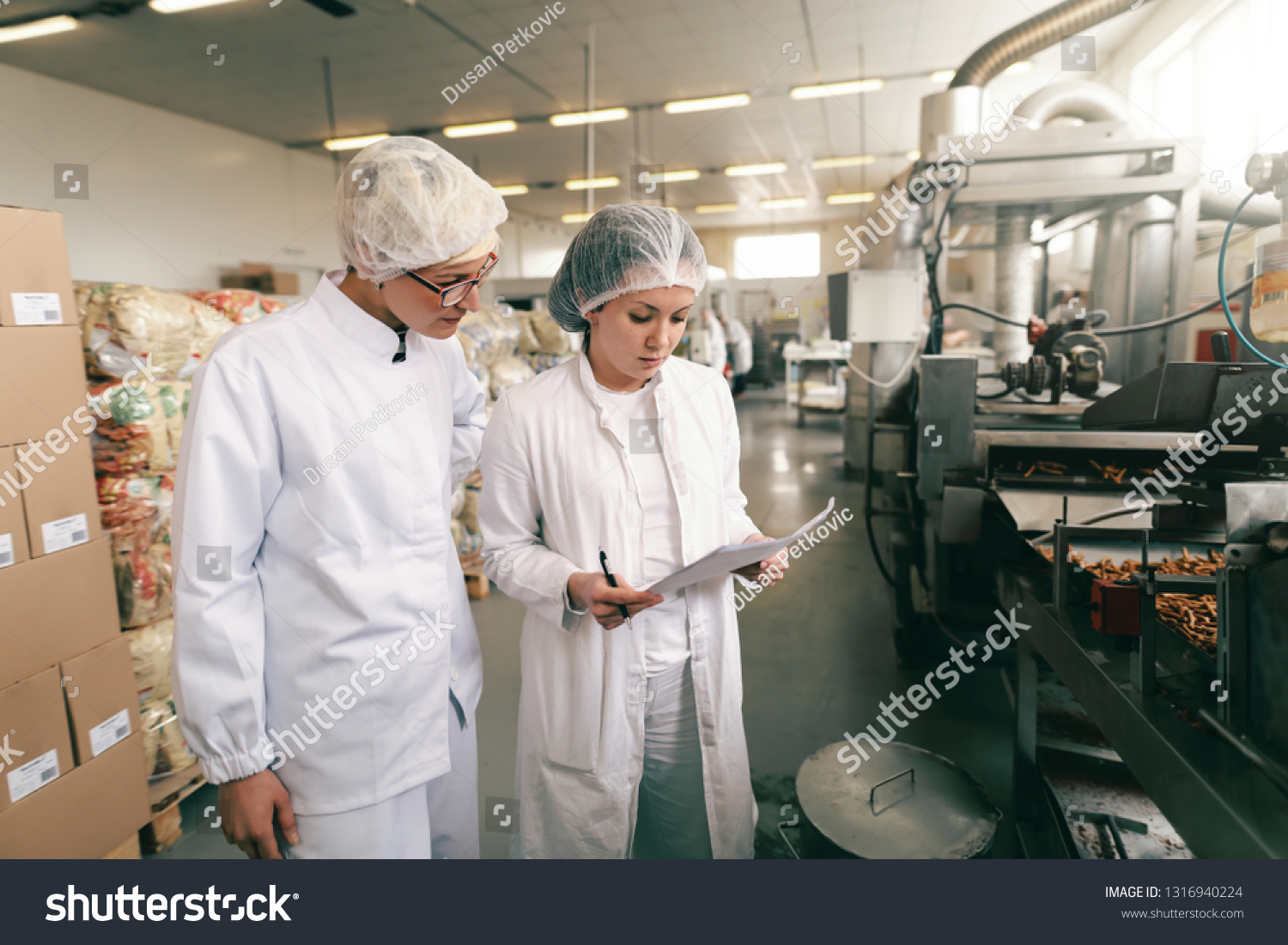 Two quality professionals in white sterile uniforms checking quality of salt sticks while standing in food factory. #1316940224