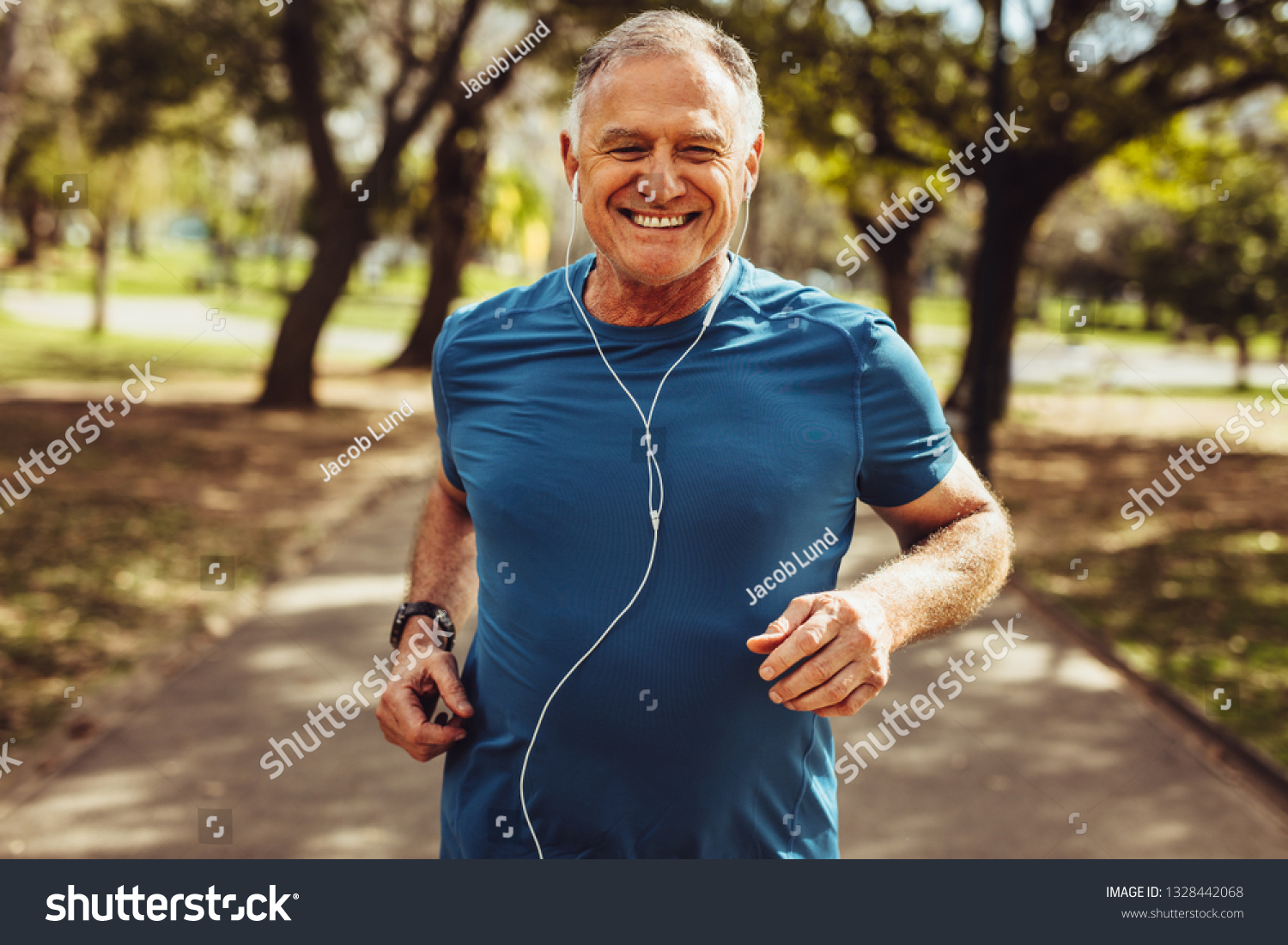 Portrait of a senior man in fitness wear running in a park. Close up of a smiling man running while listening to music using earphones. #1328442068