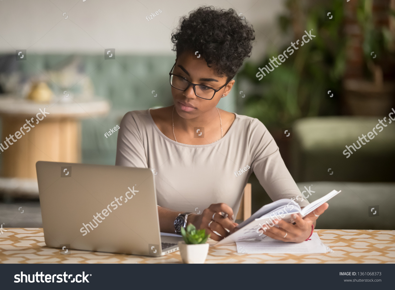 Focused young african american businesswoman or student looking at laptop holding book learning, serious black woman working or studying with computer doing research or preparing for exam online #1361068373
