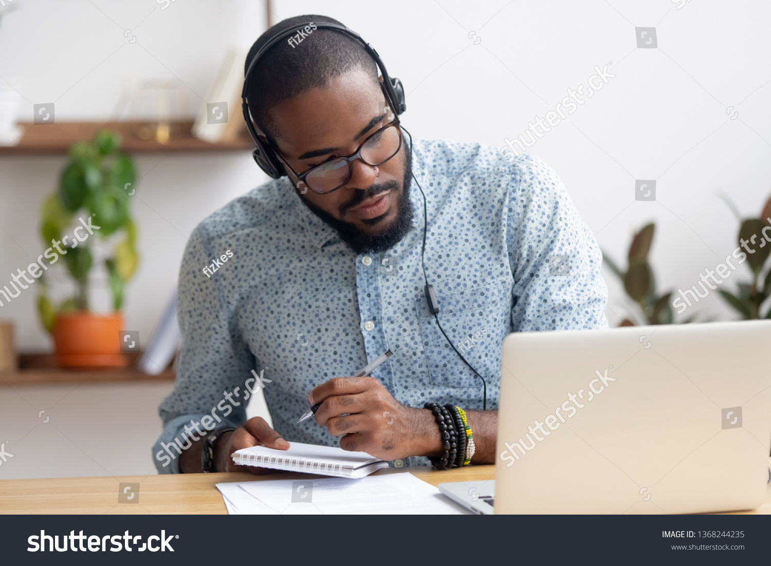 Focused african business man in headphones writing notes in notebook watching webinar video course, serious black male student looking at laptop listening lecture study online on computer e learning #1368244235