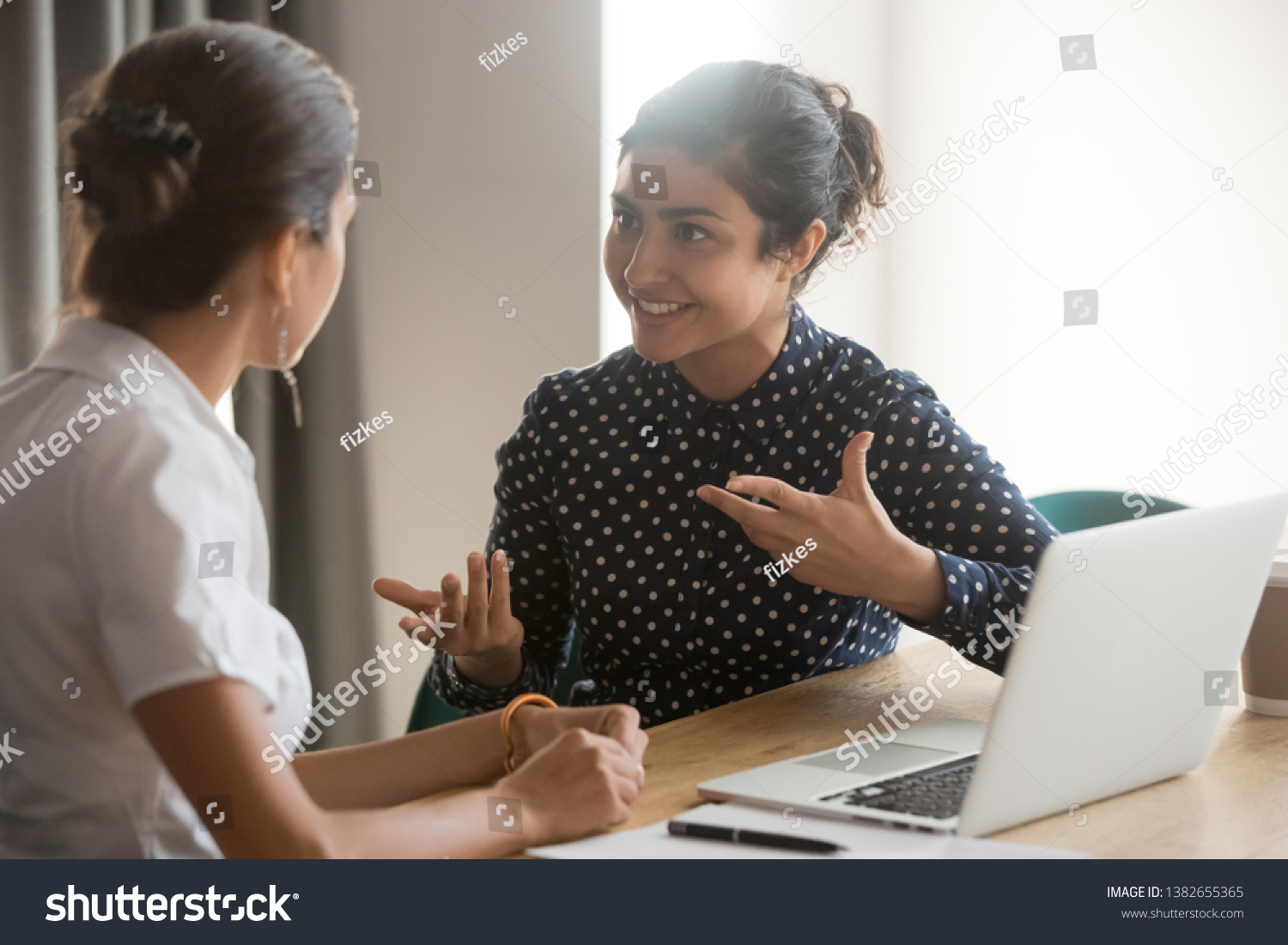 Excited multiethnic female employees discuss work issues sitting at office table, smiling diverse women workers or colleagues engaged in brainstorming talk chatting, explain ideas at workplace #1382655365