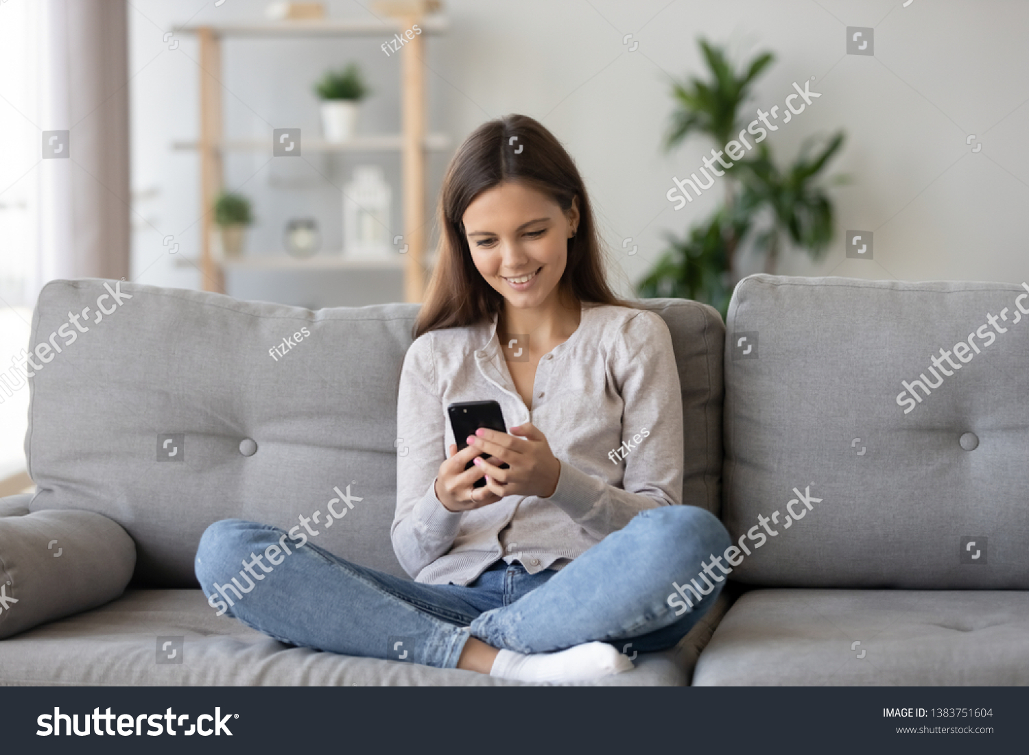 Smiling teenager girl relax sit on sofa at home using cellphone texting chatting with friend, happy young woman on couch hold smartphone shop online or check mobile application or social media #1383751604