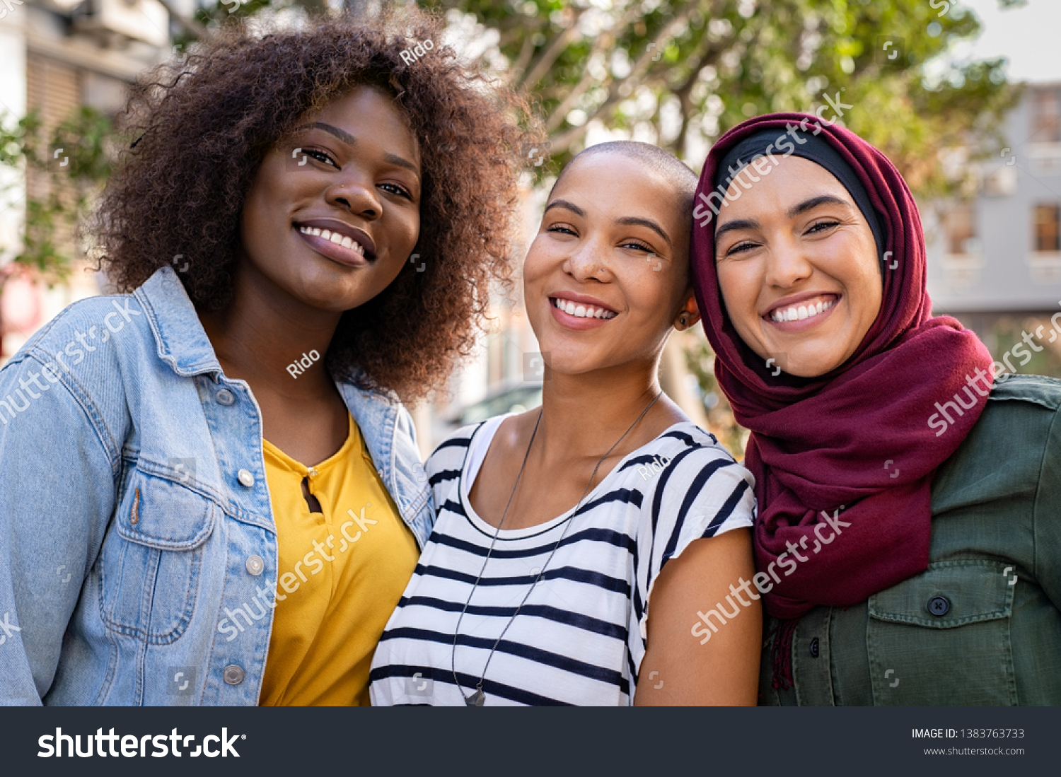 Group of three happy multiethnic friends looking at camera. Portrait of young women of different cultures enjoying vacation together. Smiling islamic girl with two african american friends outdoor. #1383763733