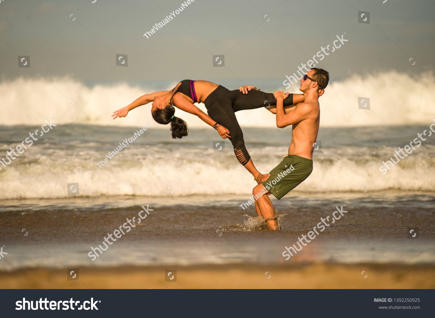young attractive and concentrated couple of acrobats practicing acro yoga balance and meditation exercise on beautiful beach under a blue sky in mind and body control and healthy lifestyle #1392250925