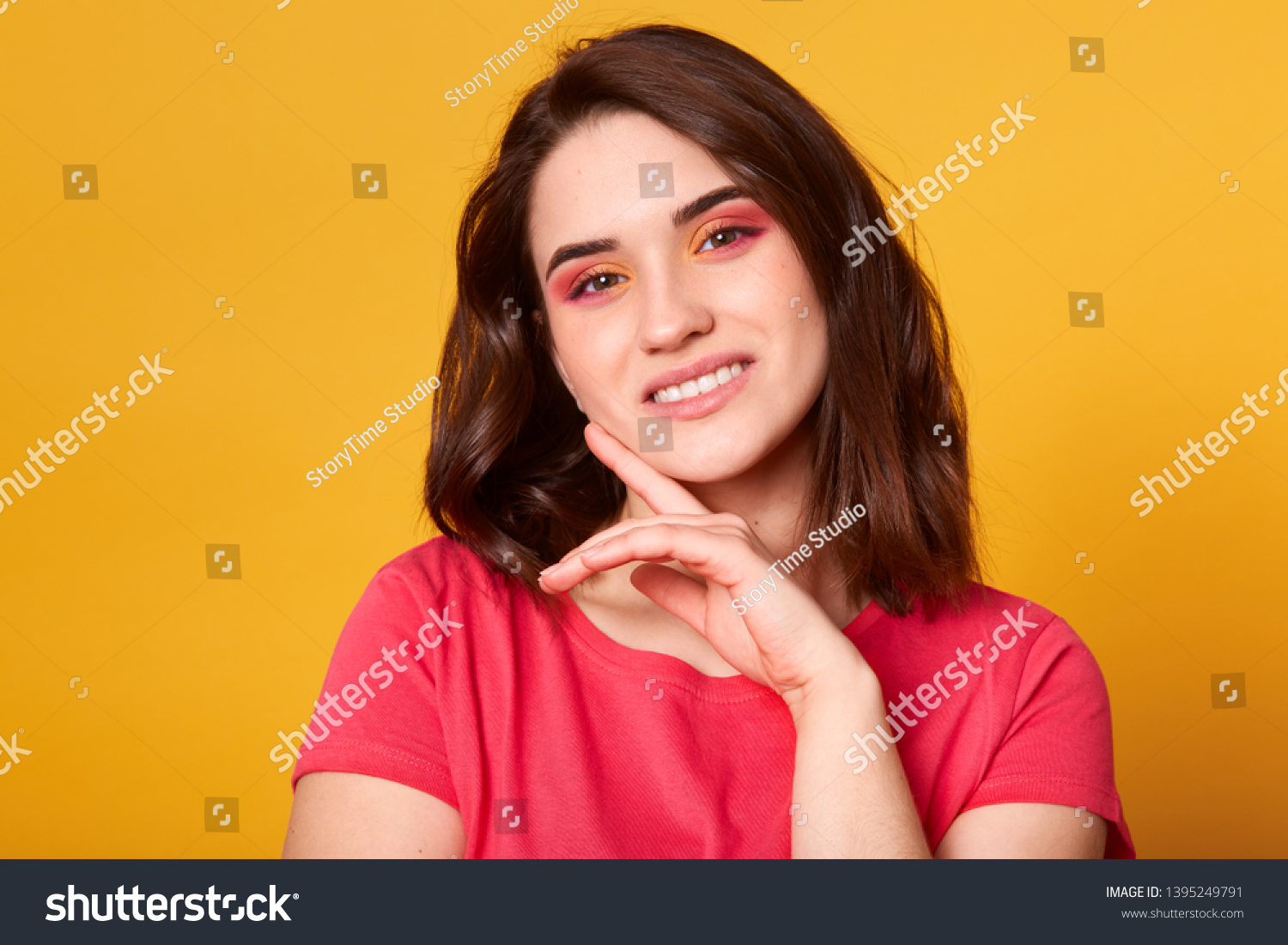 Indoor shot of beautiful brunette female with pink and orange colors makeup isolated over yellow background, looking at camera with pleasant smile, wearing casual red t shirt. Beauty concept. #1395249791