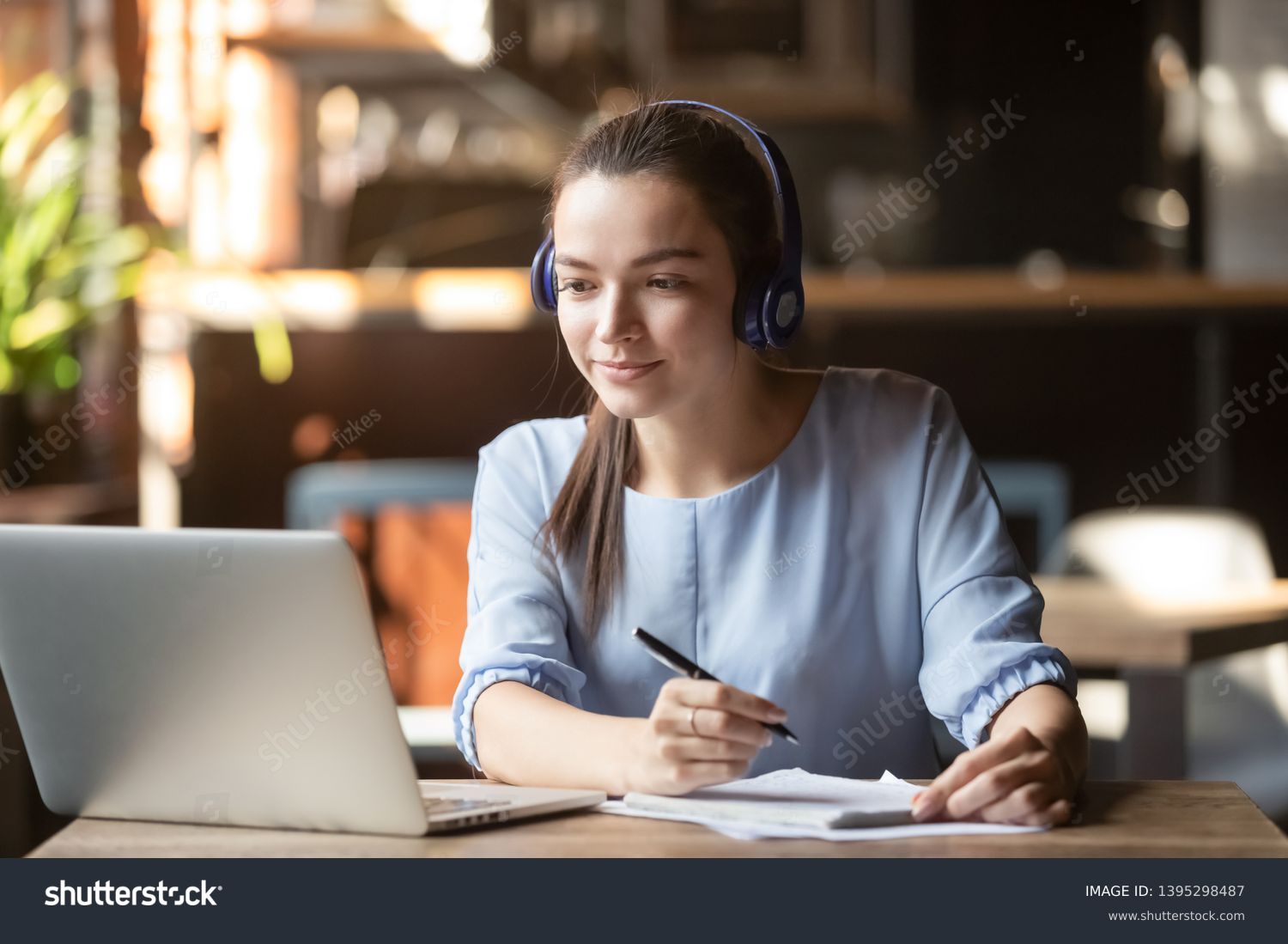 Focused woman wearing headphones using laptop in cafe, writing notes, attractive female student learning language, watching online webinar, listening audio course, e-learning education concept #1395298487