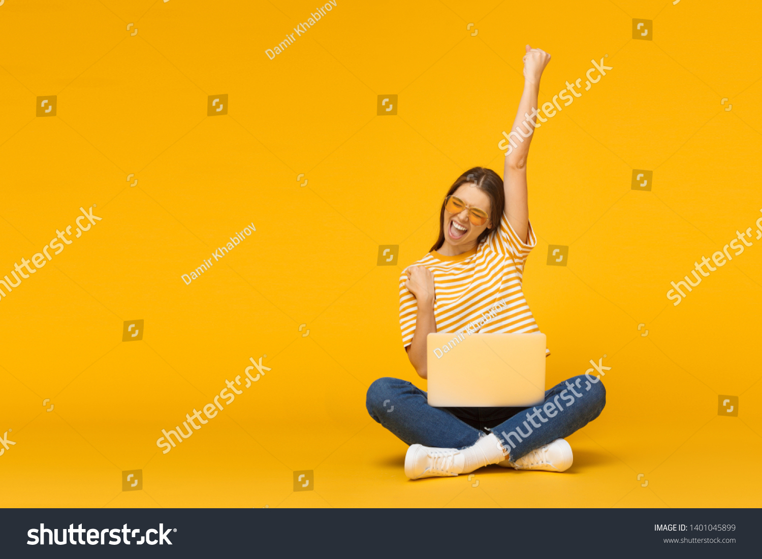 Winner! Excited smiling girl sitting on floor with laptop, raising one hand in the air is she wins, isolated on yellow background #1401045899