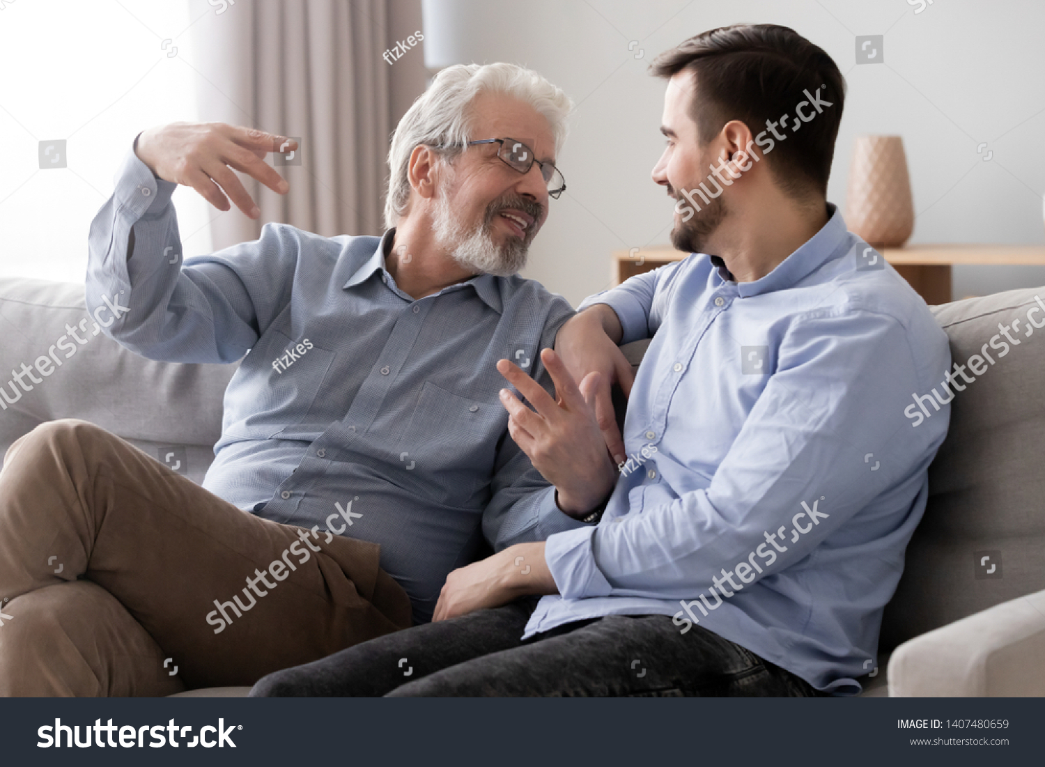 Smiling senior father sit on couch talk chat with millennial son spend time at home together, young man relax with elderly dad speak share thoughts have fun resting together on sofa in living room #1407480659