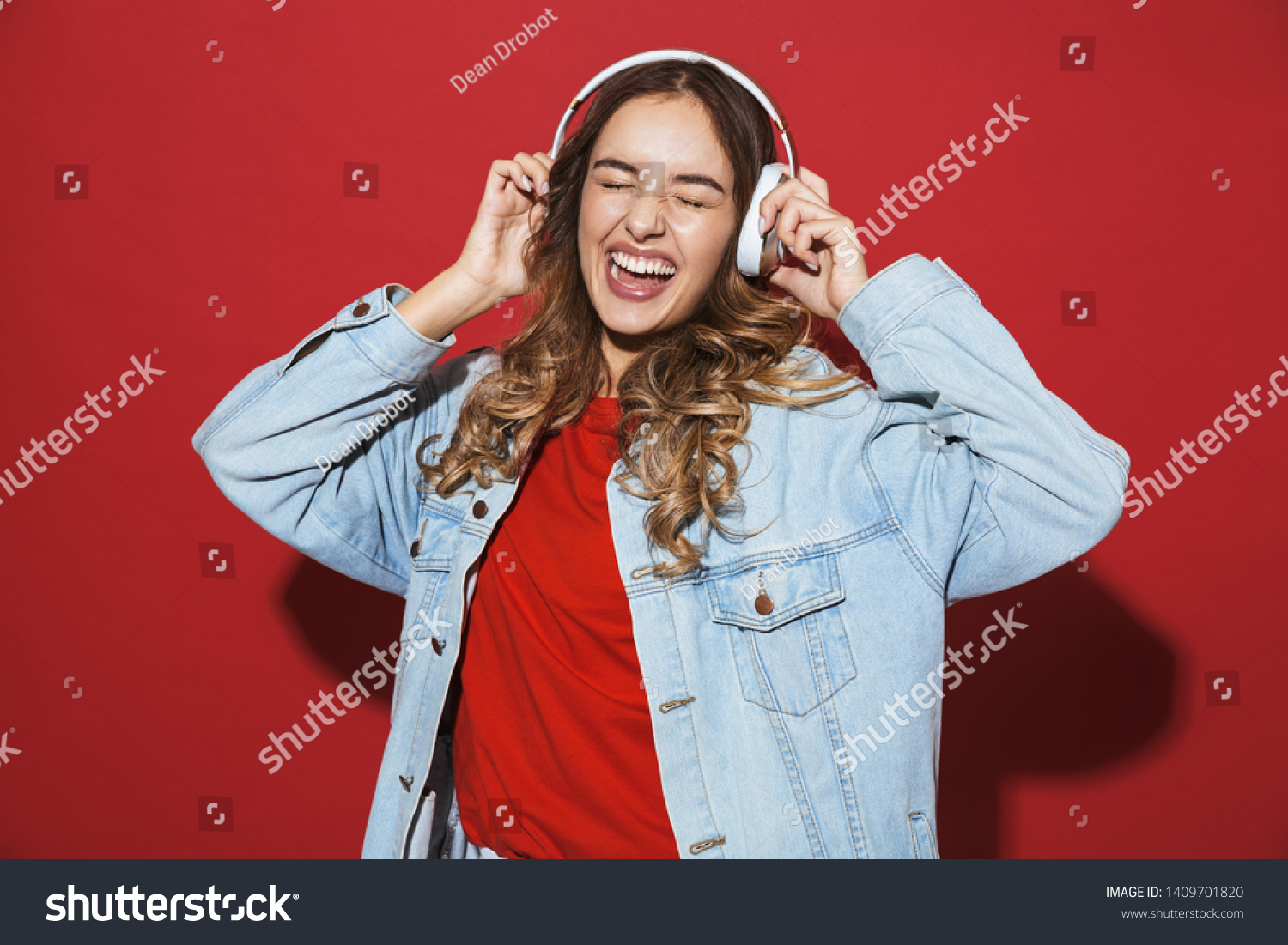 Portrait of a cheerful stylish young woman wearing denim jacket standing isolated over red background, listening to music with headphones, dancing #1409701820