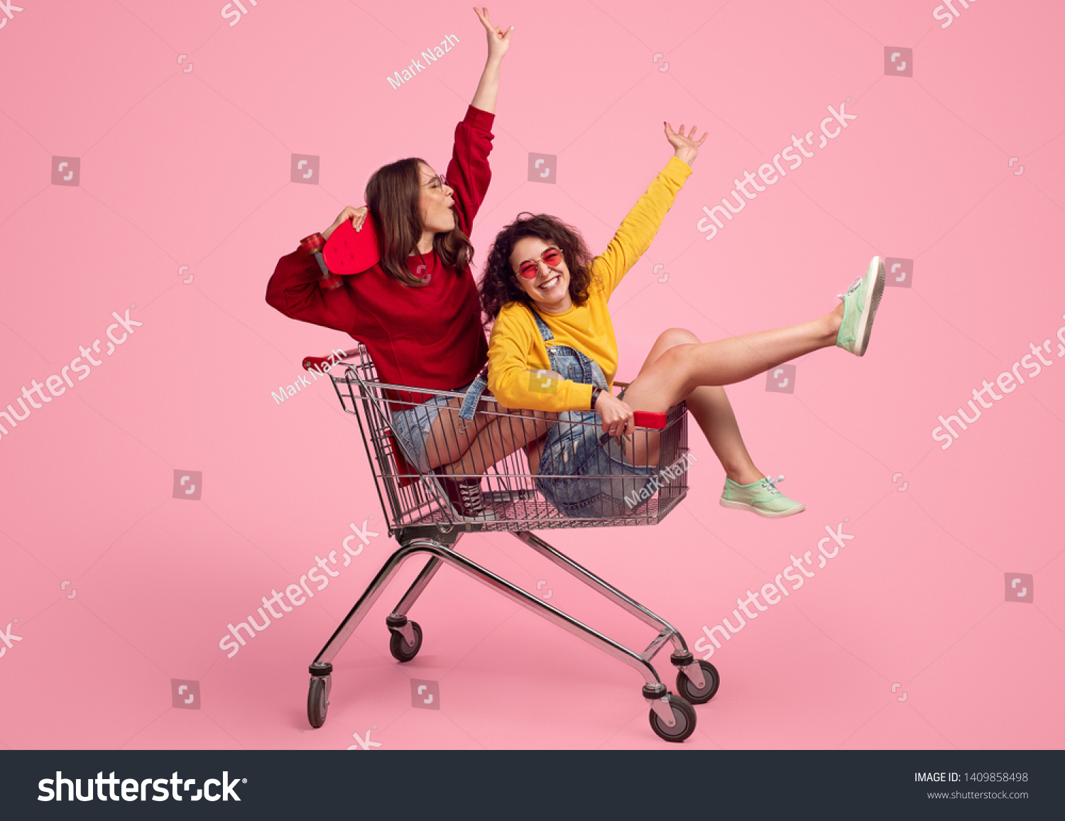 Side view of excited young friends smiling and raising hands while riding shopping trolley against pink background #1409858498