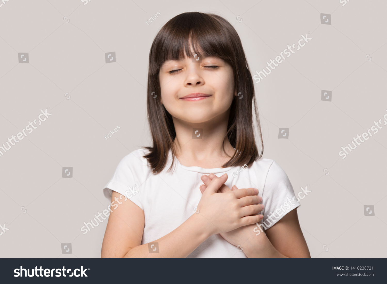 Little sincere adorable girl closed eyes holding hands on chest feeling gratitude pose isolated on sandy color beige background, arms on heart gesture of love appreciation gratitude, adoption concept #1410238721