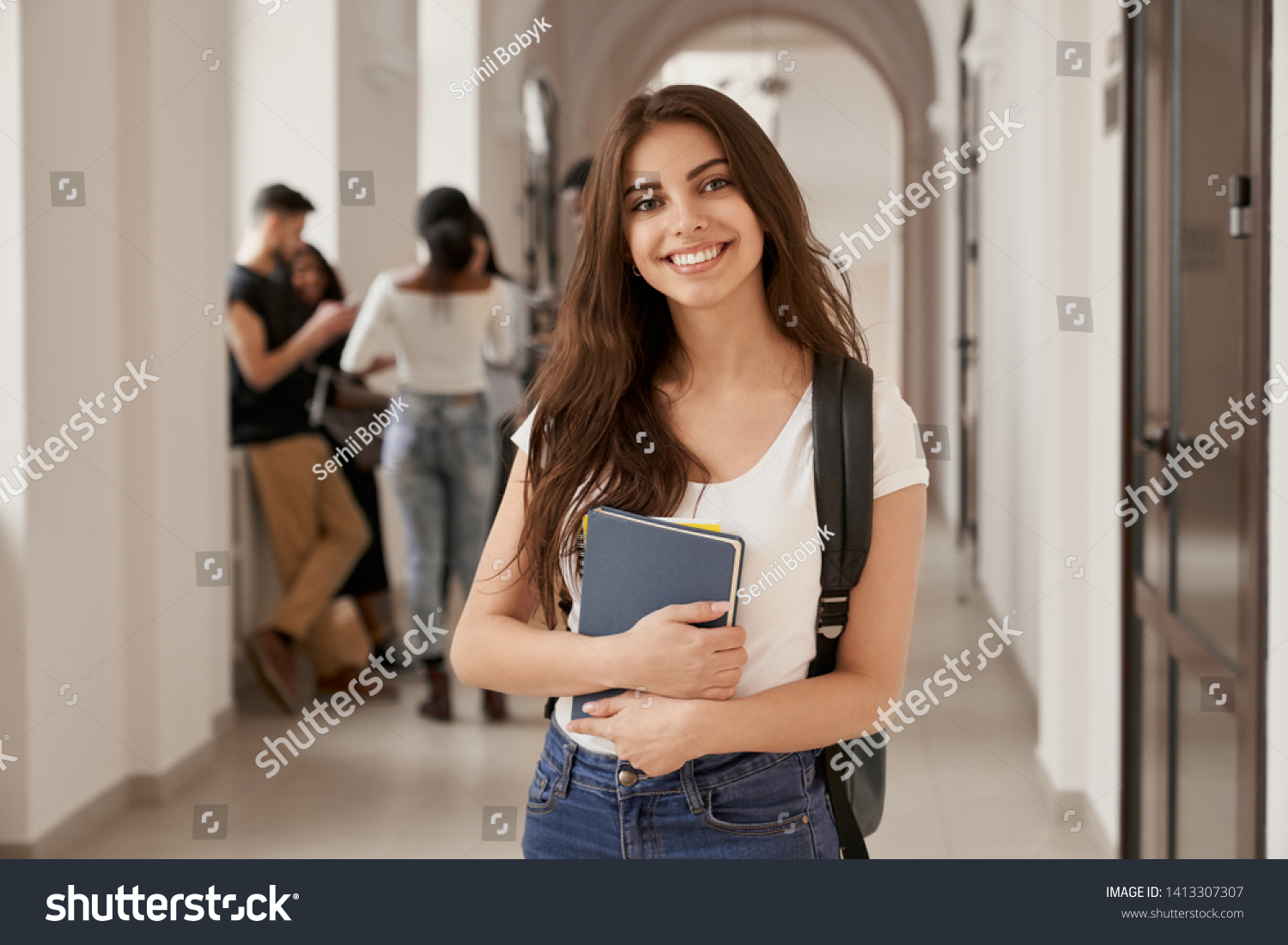 Positivity beautiful girl smiling at camera, standing on corridor with notes as backpack, going to lesson. Happy brunette female student studying in luxury university. #1413307307