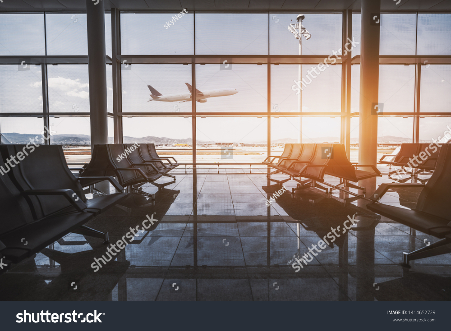 Wide-angle view of a modern aircraft gaining the altitude outside the glass window facade of a contemporary waiting hall with multiple rows of seats and reflections indoors of an airport terminal #1414652729