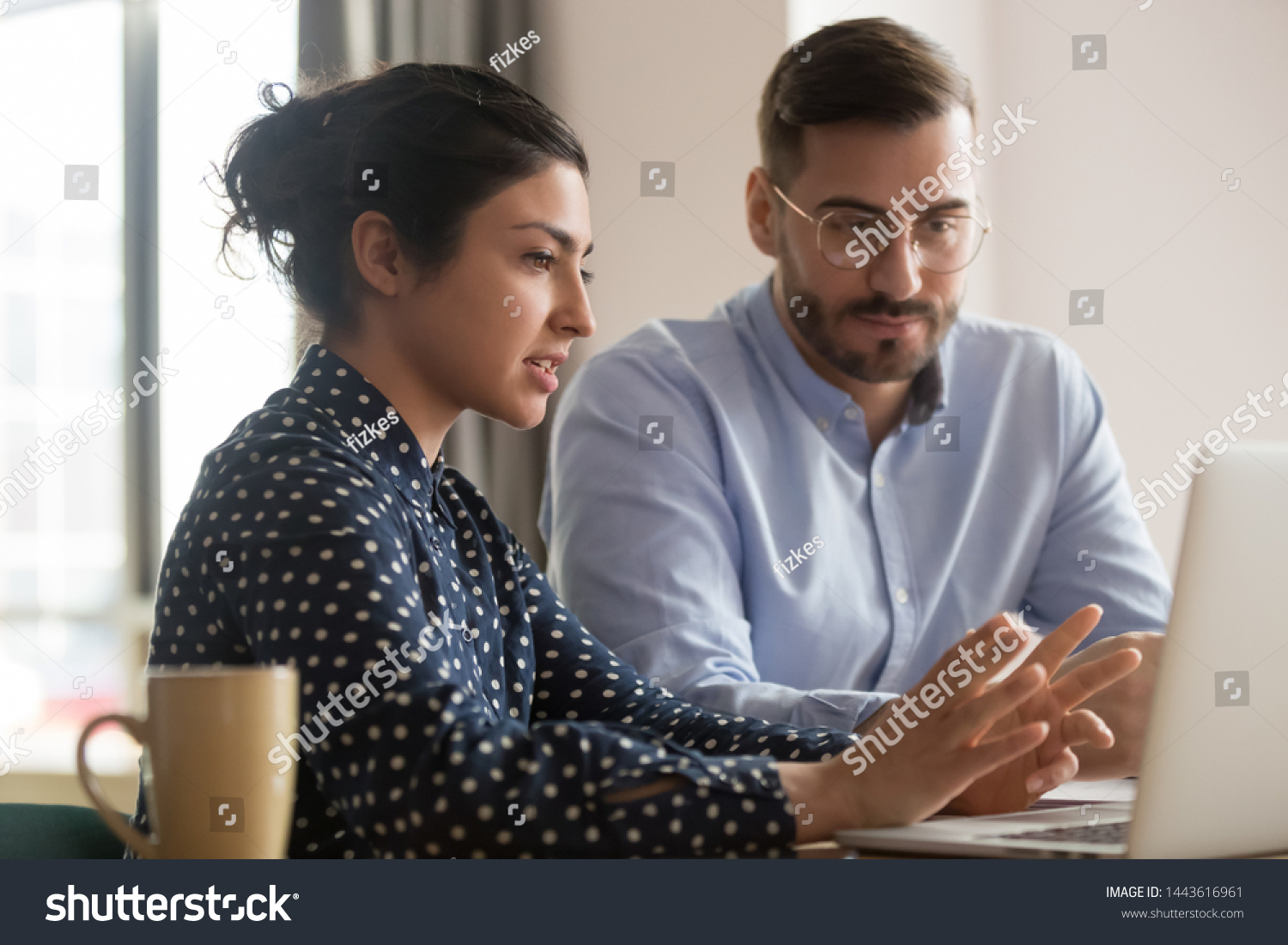 Focused male intern listening to serious indian business woman mentor teacher explaining online strategy looking at laptop computer teach trainee training new worker learning new skill at workplace #1443616961