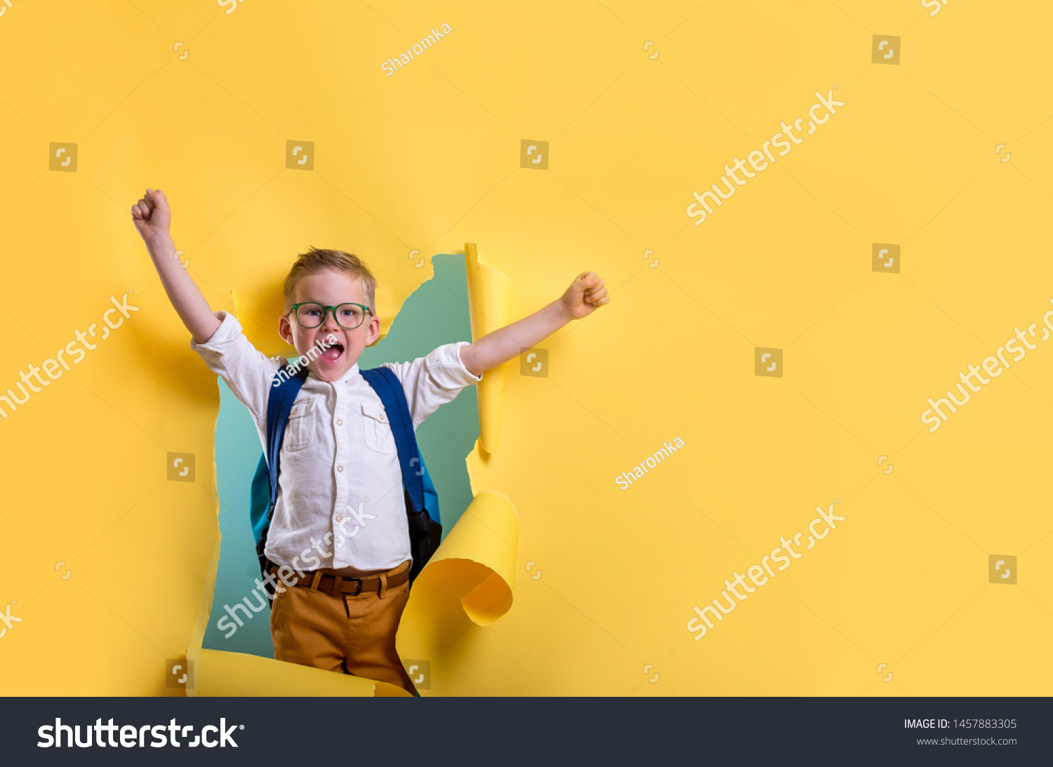 Child boy with book and bag breaking through yellow paper wall. Happy 
smiling kid go back to school, kindergarten. Success, motivation, winner, genius concept. Little kid dreaming to be superhero #1457883305
