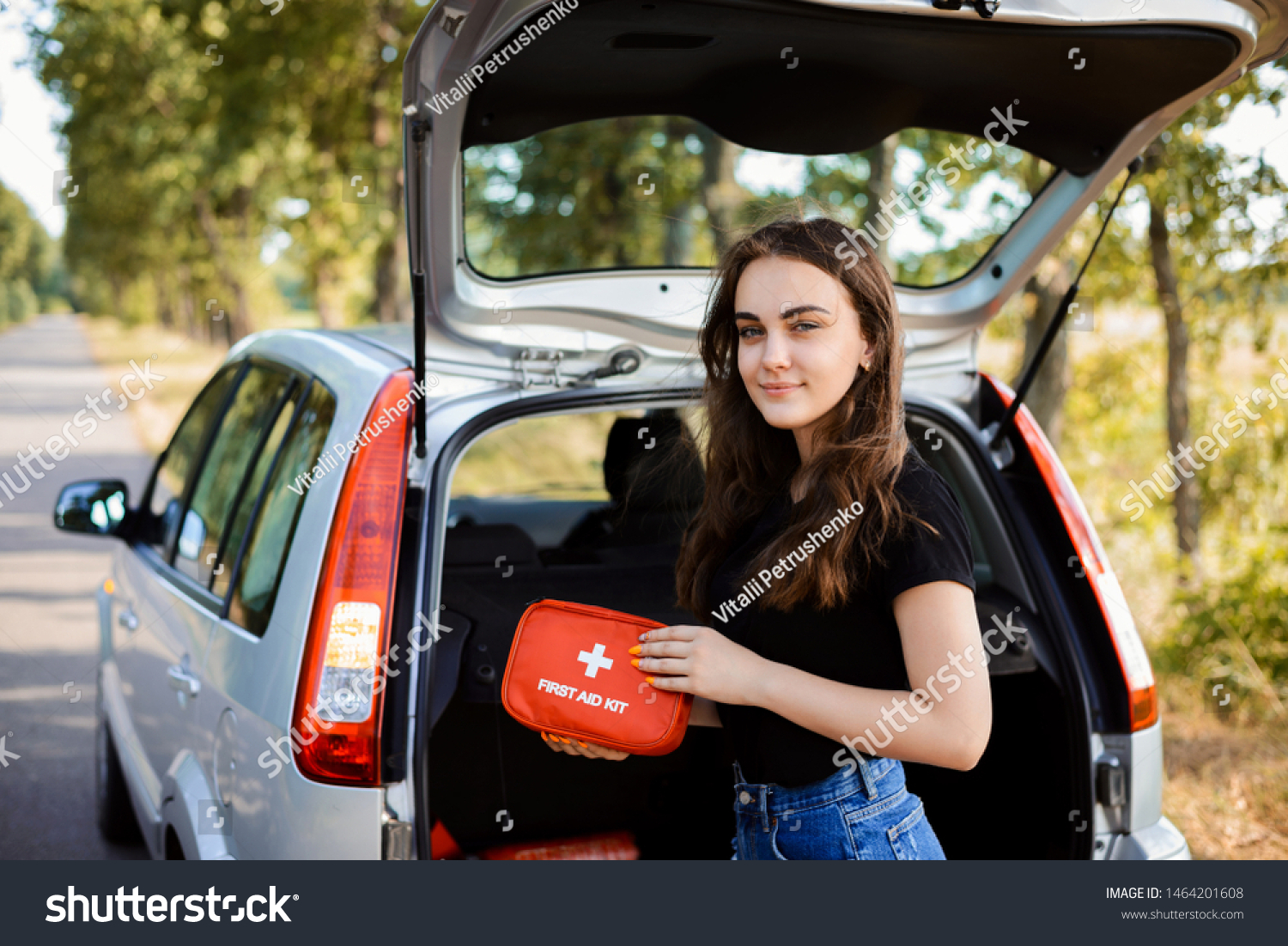 Young girl standing near open back door of silver hatchback car and shows first aid kit that must be in every car for emergency #1464201608