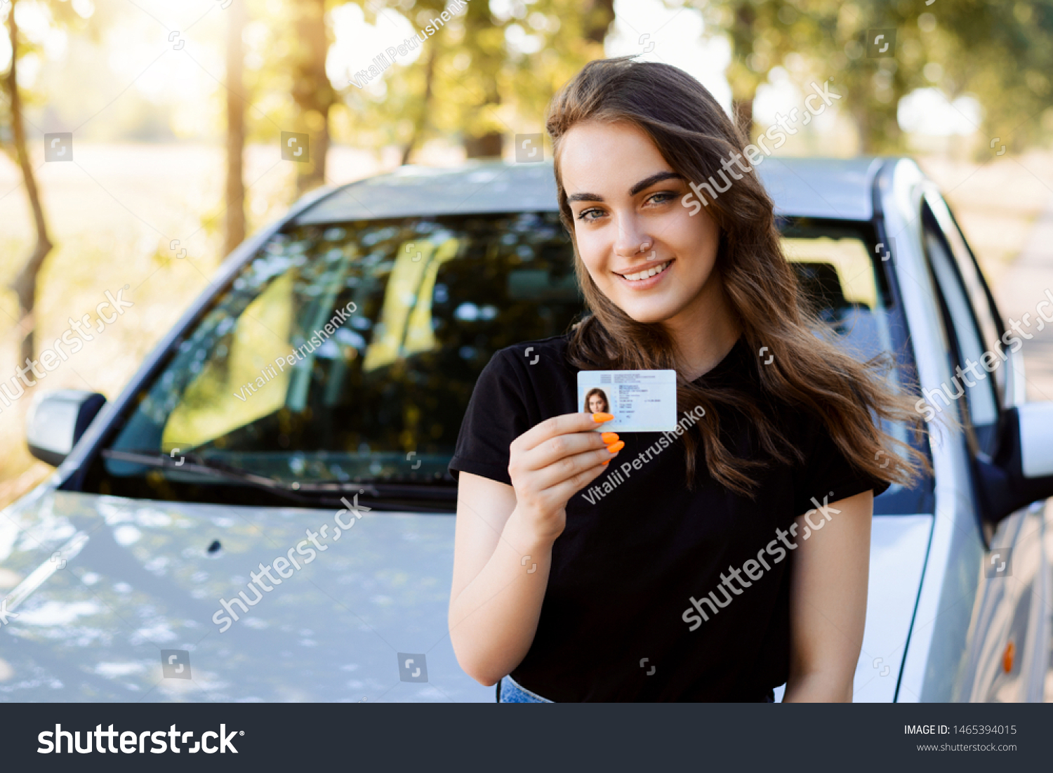 Attractive young woman wearing black T-shirt standing in front of modern car bragging about receiving driving license showing licence to the camera and feeling happy #1465394015